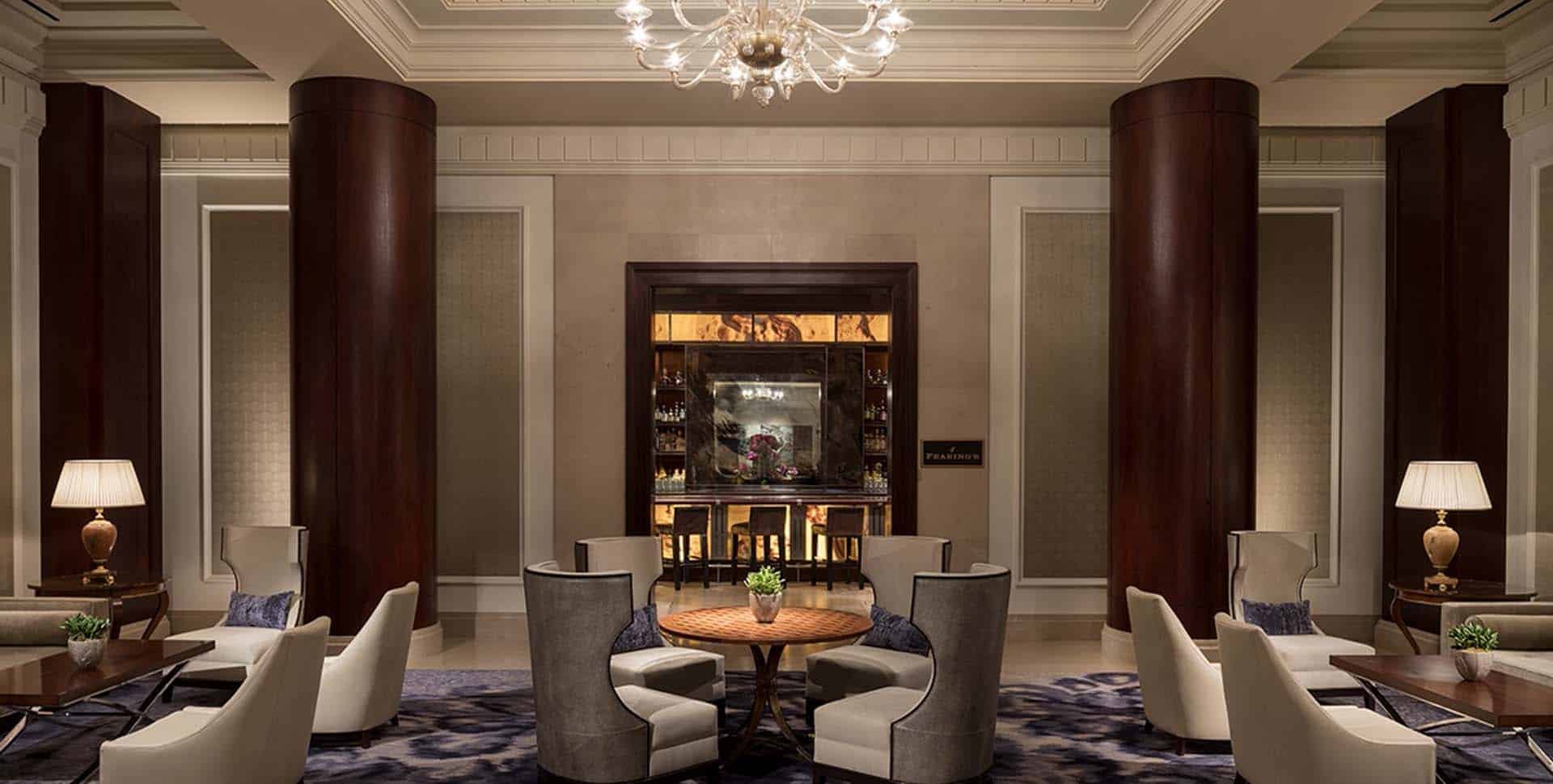 A hotel lounge with armchairs around a round wooden table, two large columns, decorative lamps, and a chandelier overhead. A bar is visible in the background.