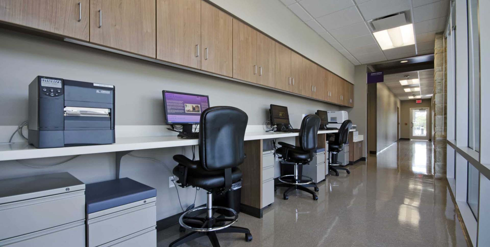 A row of computer workstations with office chairs and printers along a hallway with wooden cabinets and overhead lighting.