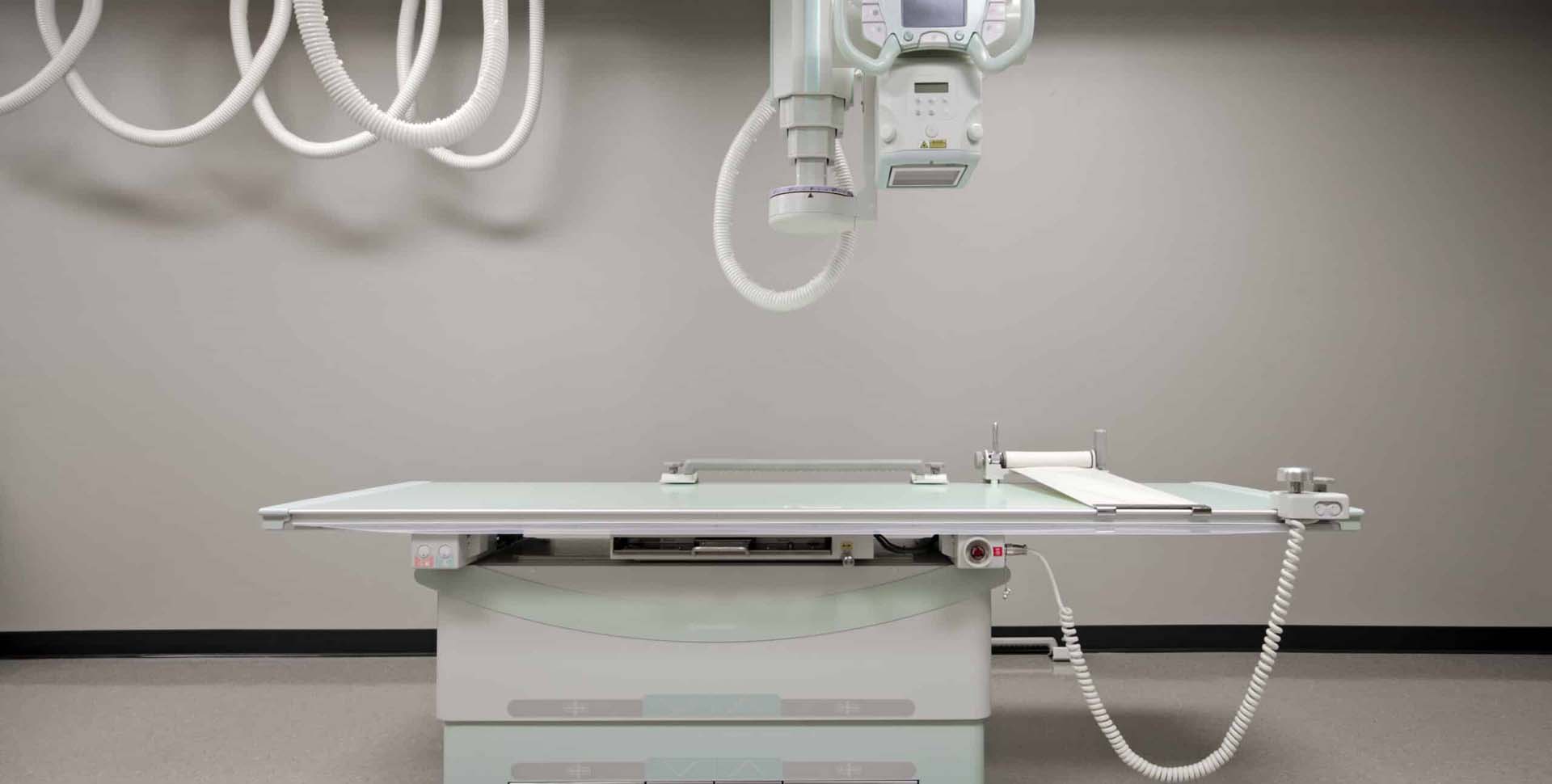 A medical X-ray machine is positioned above an empty examination table in a clean, modern imaging room.