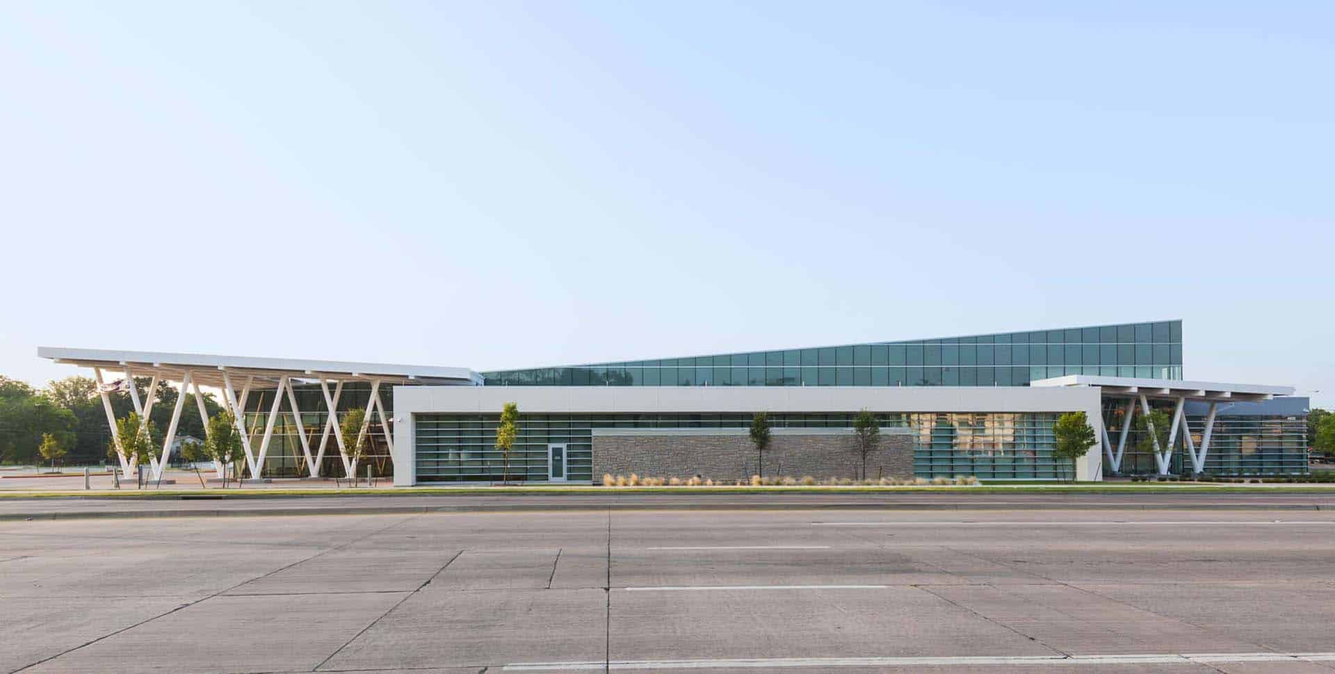 Modern building with glass walls and angular rooflines, featuring white support beams, situated next to an empty road under clear sky.