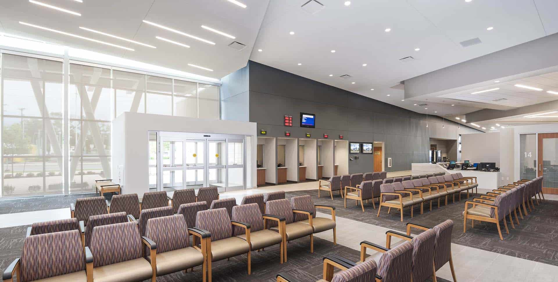 Modern waiting area with rows of empty chairs, large windows, and digital screens on the wall; reception desk visible in the background.