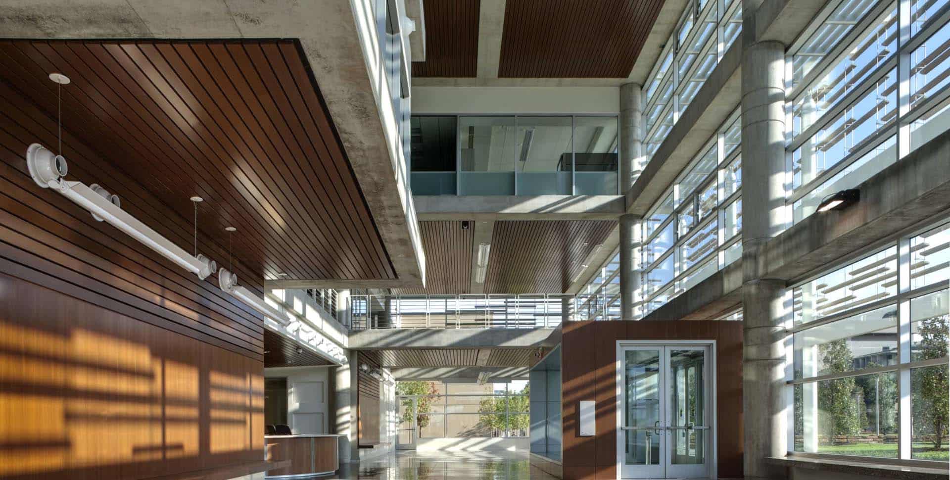 Modern building interior with large windows, concrete pillars, wooden panels, and natural light streaming into a spacious, open lobby area.