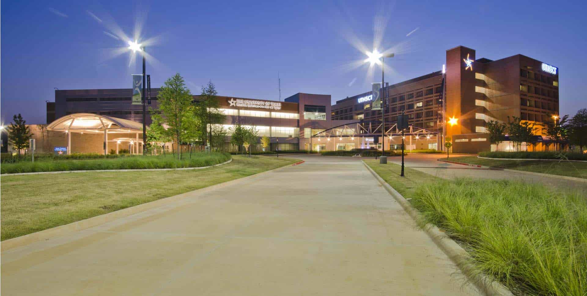 A modern hospital complex with illuminated signs, landscaped greenery, and a wide driveway leading to the main entrance at dusk.