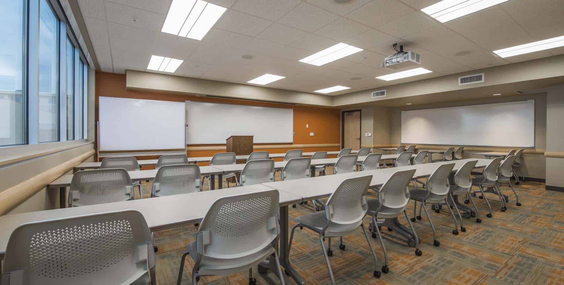 Empty classroom with rows of gray chairs and desks facing a whiteboard, windows on the left, and a ceiling-mounted projector.
