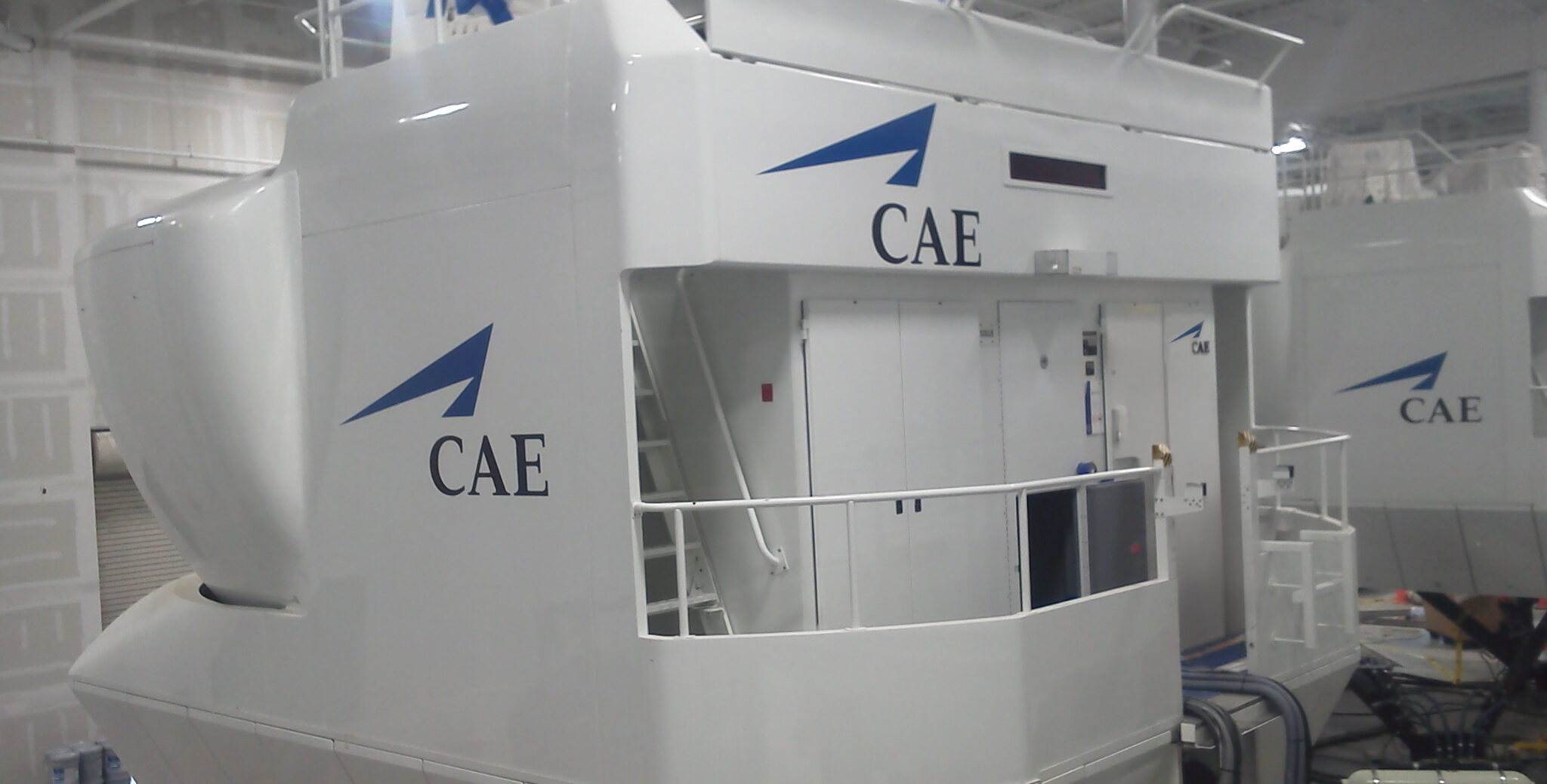 A white CAE flight simulator with a blue logo is positioned indoors, featuring exterior stairs, railings, and a red ladder near the entrance.