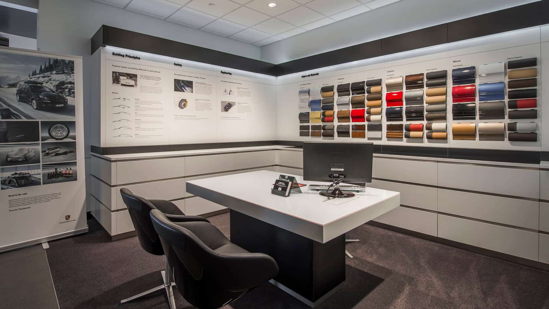 A modern car showroom office with a desk, two chairs, a computer, and wall displays featuring color and material samples for car customization.