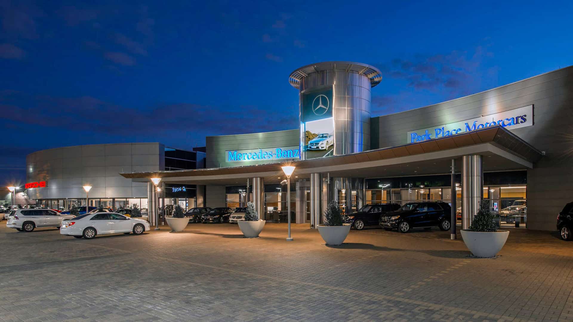 Exterior view of a Mercedes-Benz dealership at dusk, with several cars parked outside and illuminated signage on the building.