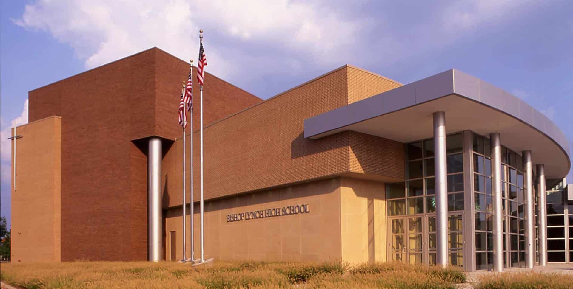 Modern brick and glass school building with three flagpoles, an American flag, and a cross on the corner wall; landscaped with tall grasses.