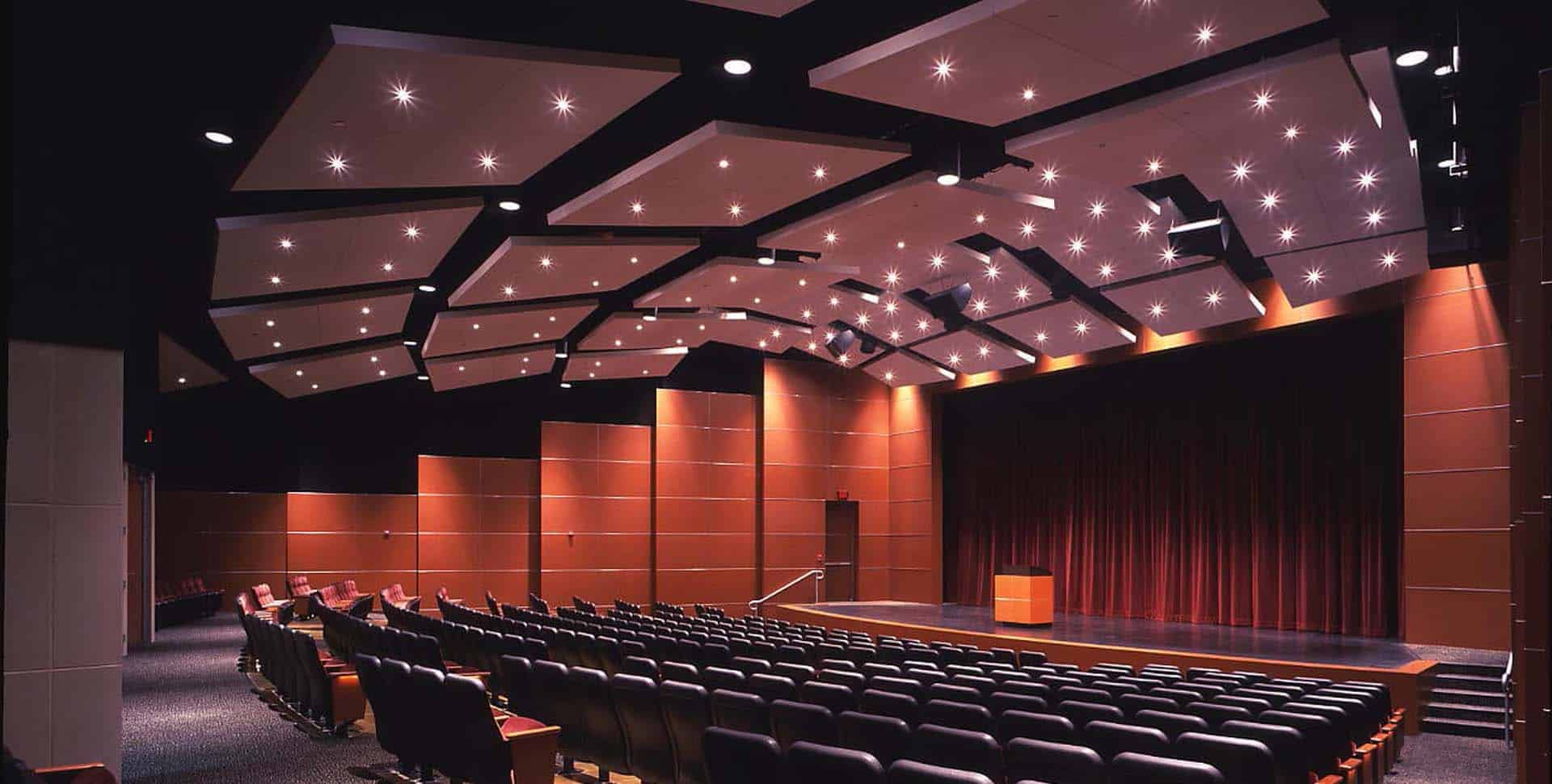 A modern auditorium with rows of empty seats, a wooden podium on stage, dark curtains, and a ceiling with geometric panels and recessed lights.