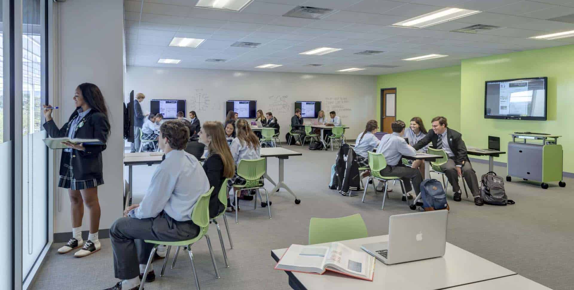 Students in uniforms work in groups and individually in a bright classroom with green chairs and digital screens; one student writes on a window.