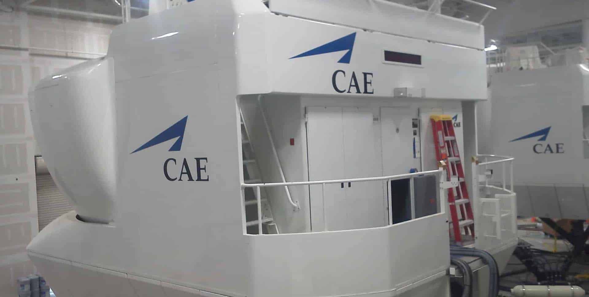 A white CAE flight simulator with a blue logo is positioned indoors, featuring exterior stairs, railings, and a red ladder near the entrance.