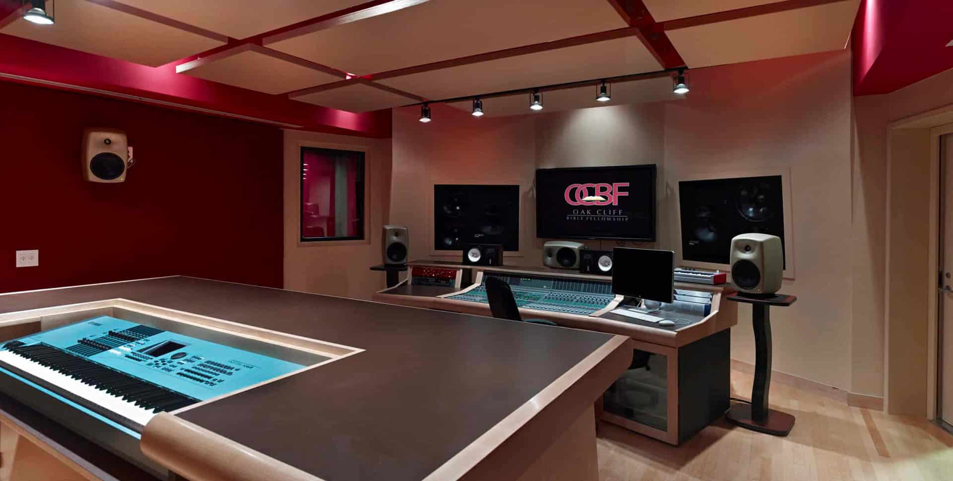 Modern recording studio with a keyboard, mixing console, large monitors, speakers, and a screen displaying the Oak Cliff Productions logo. The room has red accents and professional lighting.