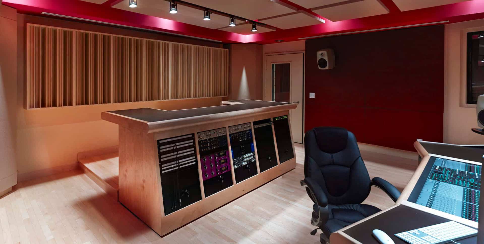 A modern recording studio with a large mixing console, audio equipment racks, an office chair, wood floors, and acoustic paneling on the walls and ceiling.