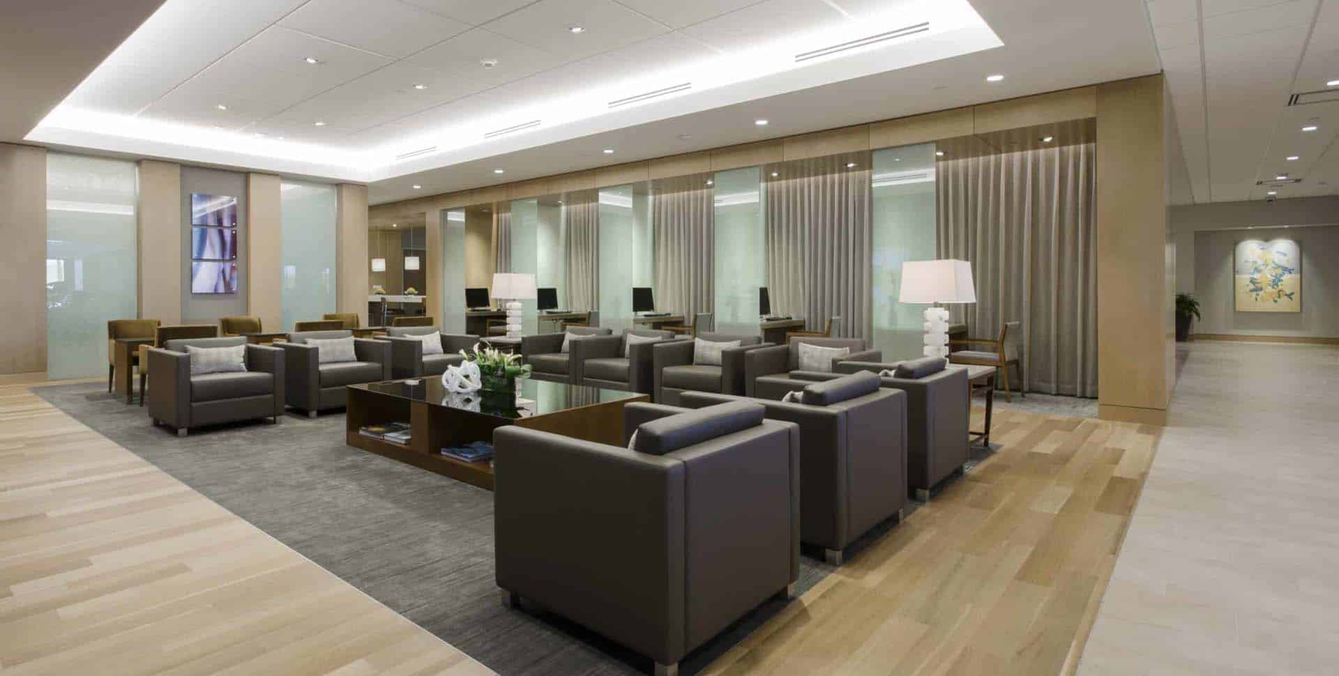 Modern lounge area with brown armchairs, tables, and decorative lamps on a wood and gray rug floor, surrounded by glass walls and neutral-toned decor.