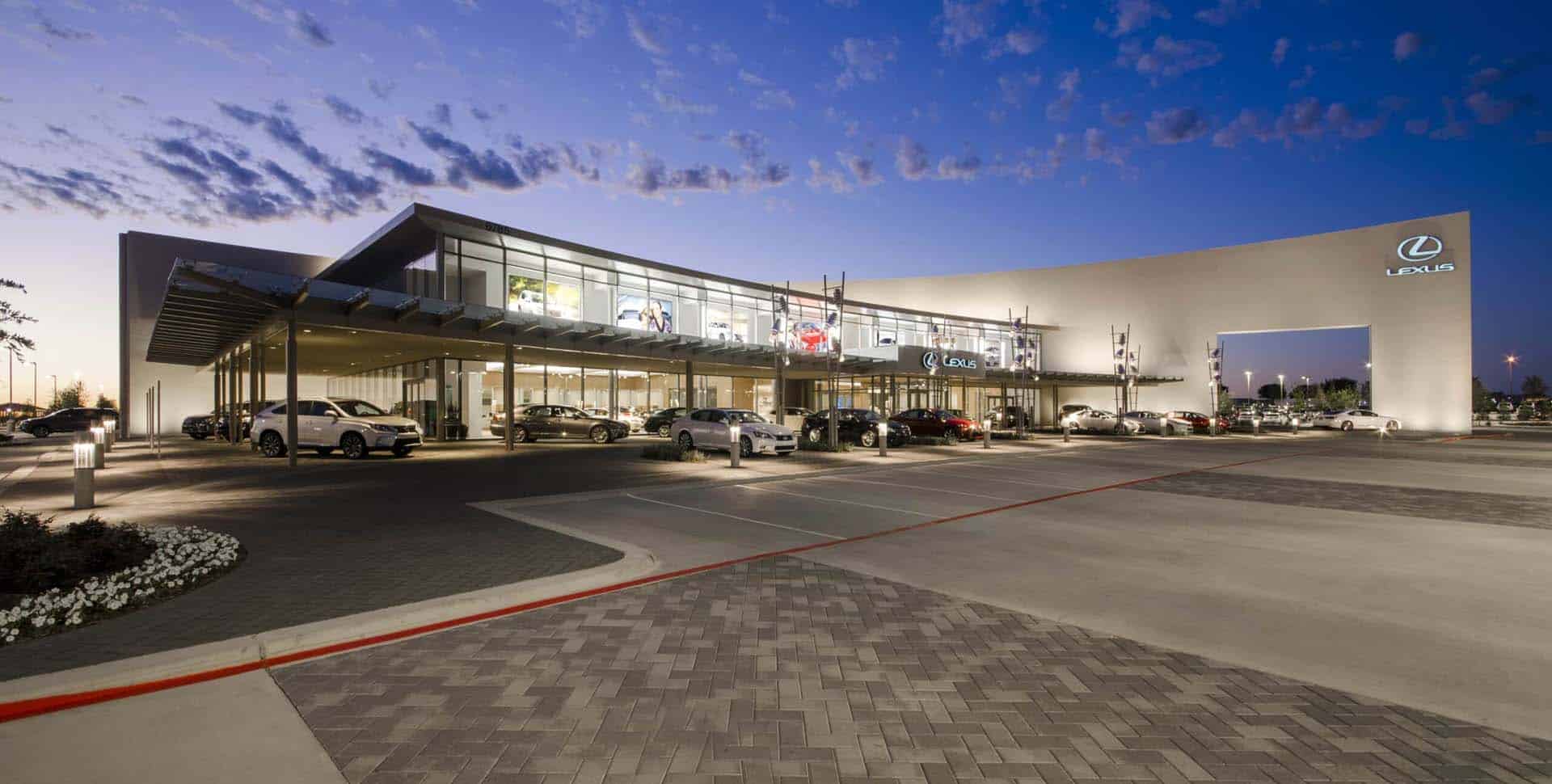 A modern Lexus dealership at dusk, featuring large glass windows, parked cars outside, and illuminated signage.