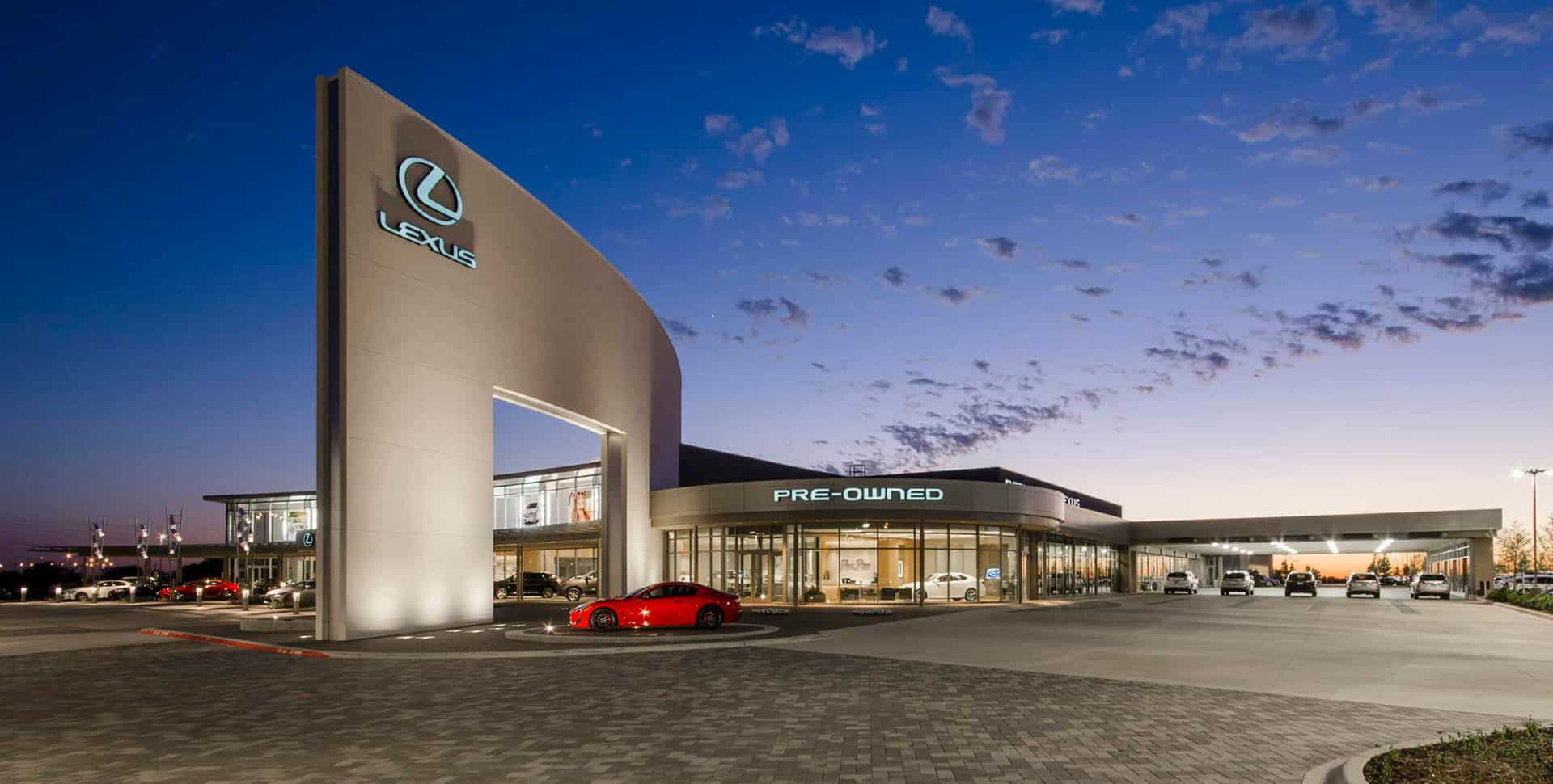 Lexus dealership at dusk with a prominent sign, modern architecture, and several cars parked in front, including a red vehicle near the entrance labeled "Pre-Owned.
