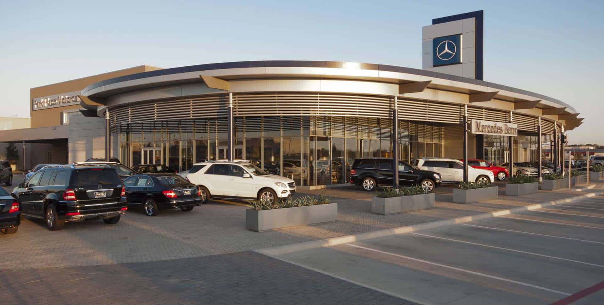 A modern Mercedes-Benz dealership building with several cars parked in front on a sunny day.