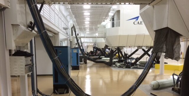 Looking out from underneath a full-flight simulator, several other full-flight simulators are lined up in a large indoor facility.