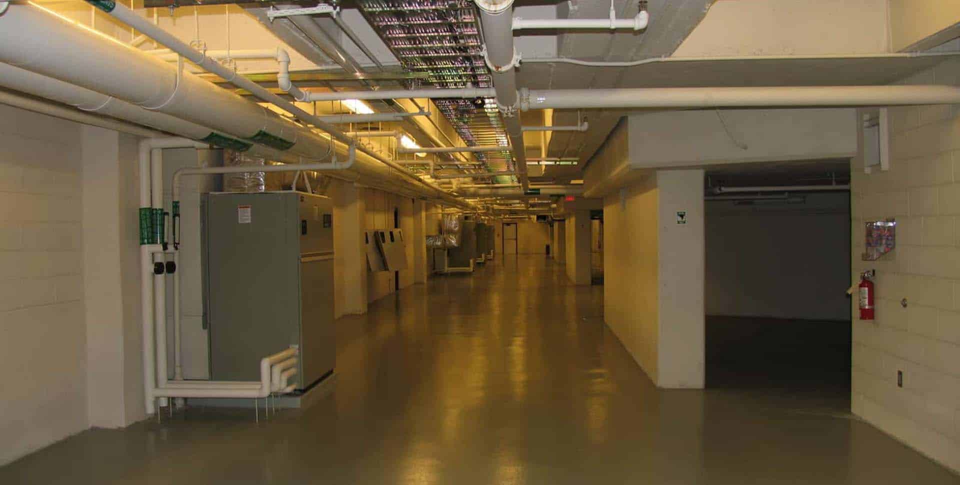 A well-lit basement hallway with exposed pipes on the ceiling, electrical panels on the left, and a fire extinguisher mounted on the right wall.