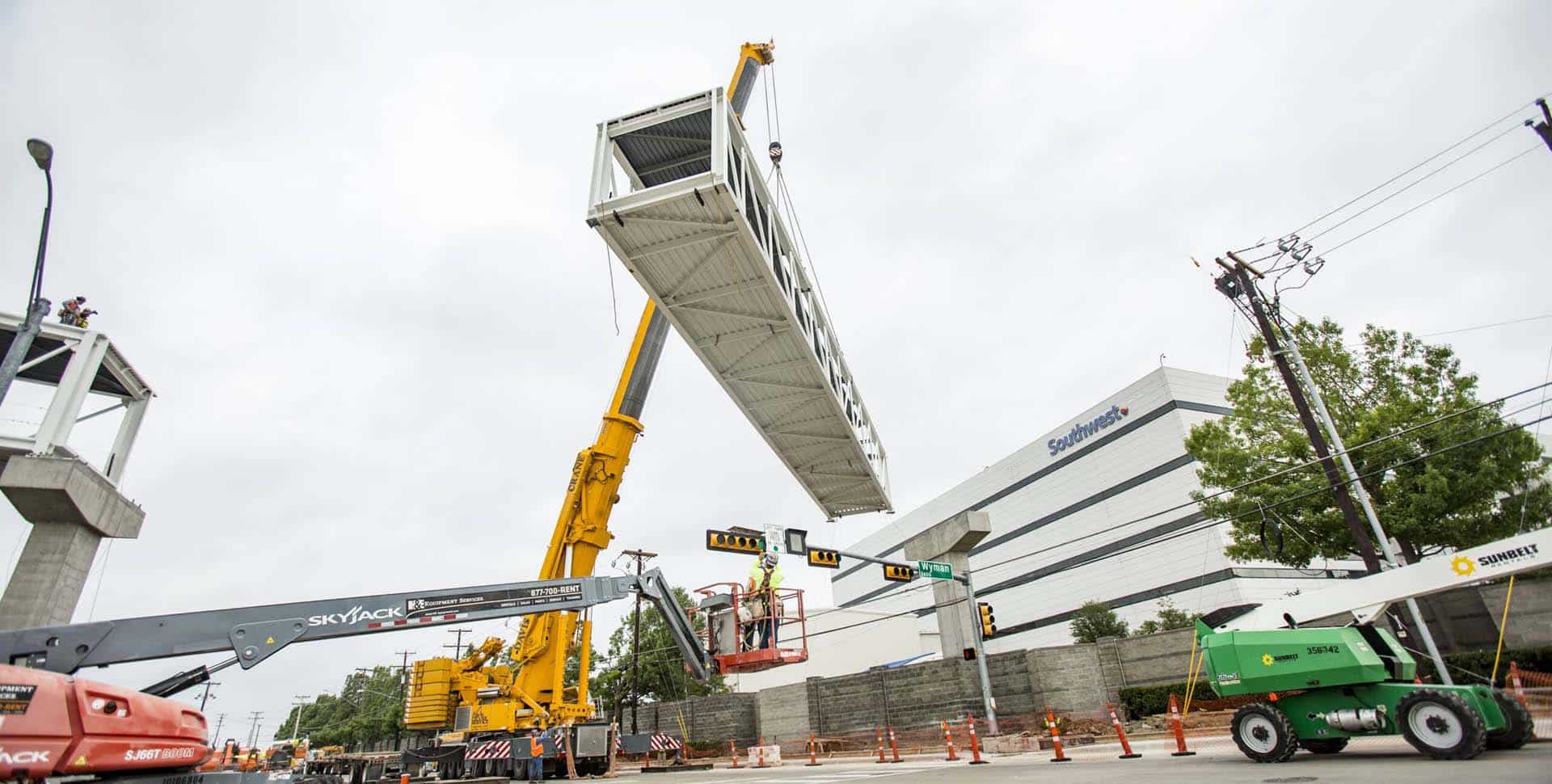 A large crane lifts a pedestrian bridge segment into place over a city street, with construction vehicles and workers present below.