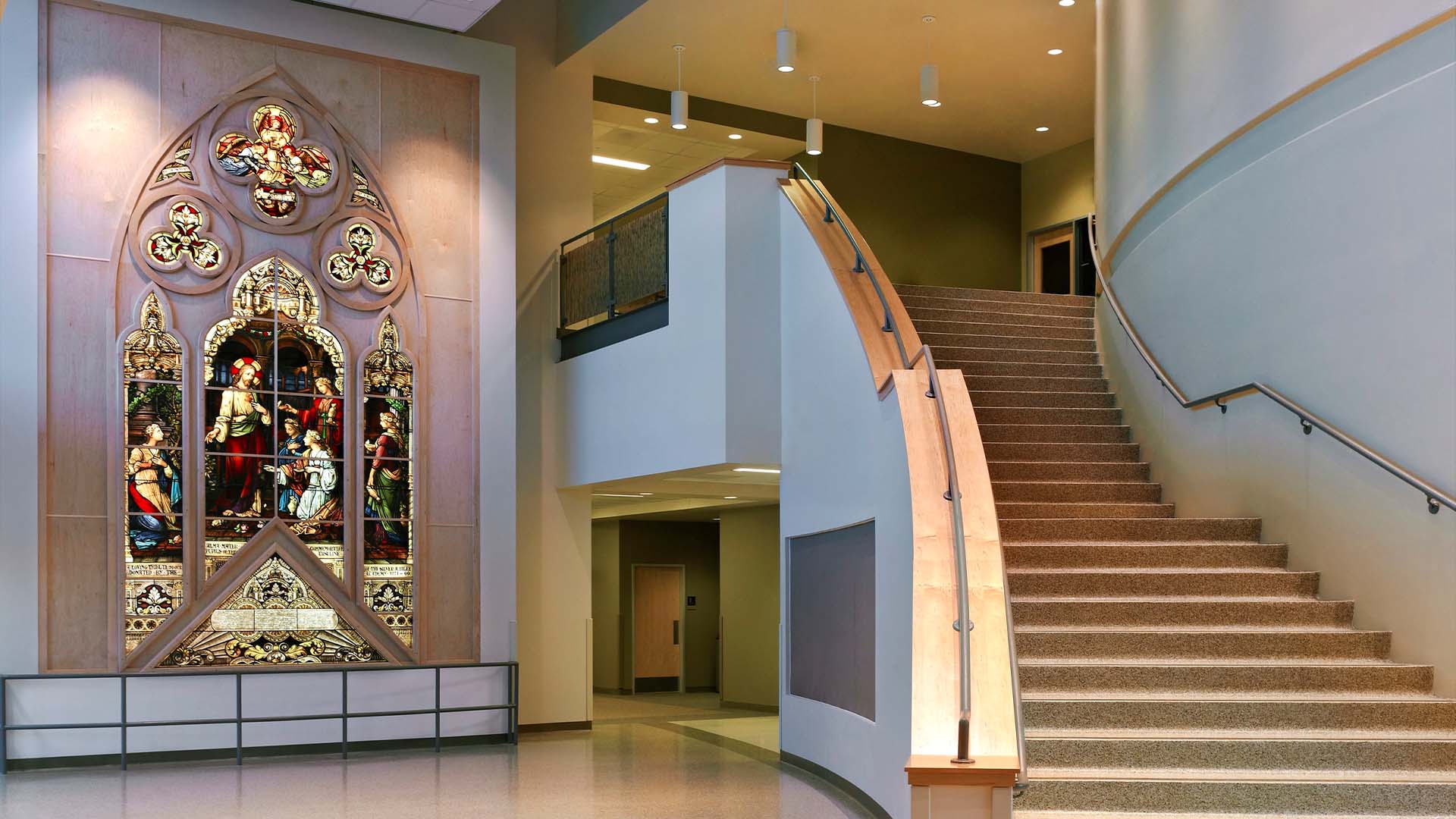 A large stained glass window is mounted on a wall next to a modern curved staircase in a spacious building interior.