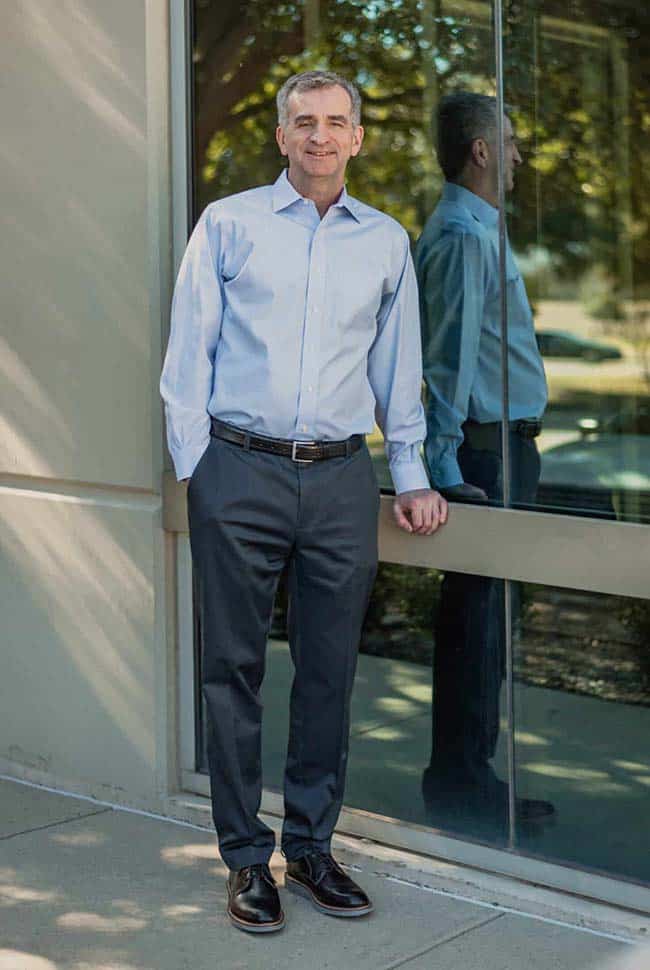 A man in a light blue shirt and dark pants stands on a sidewalk next to a glass window, with his reflection visible in the glass.