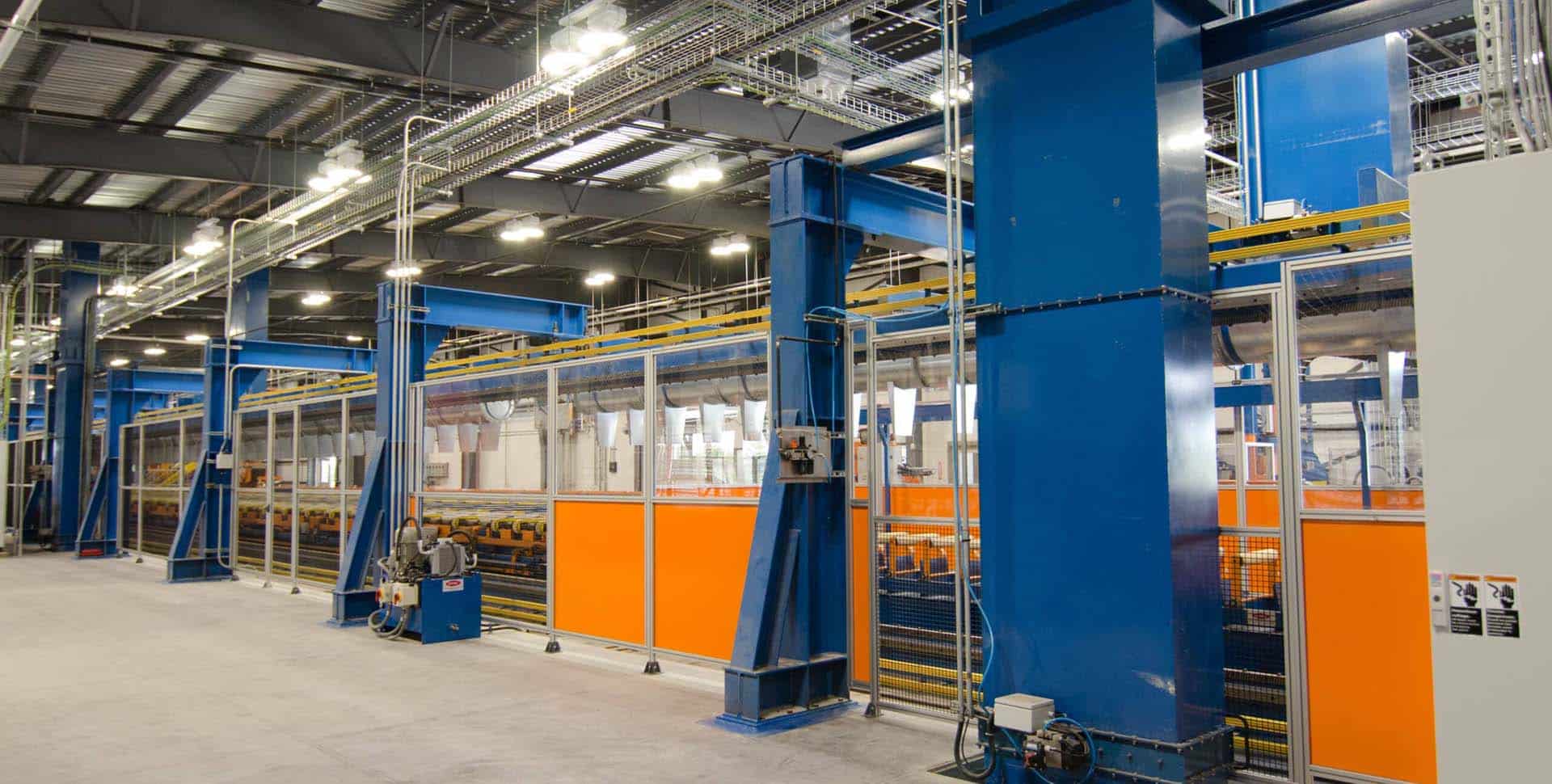 Interior view of a modern industrial facility with large blue structural supports, orange safety barriers, and overhead wiring, featuring various machinery and equipment.