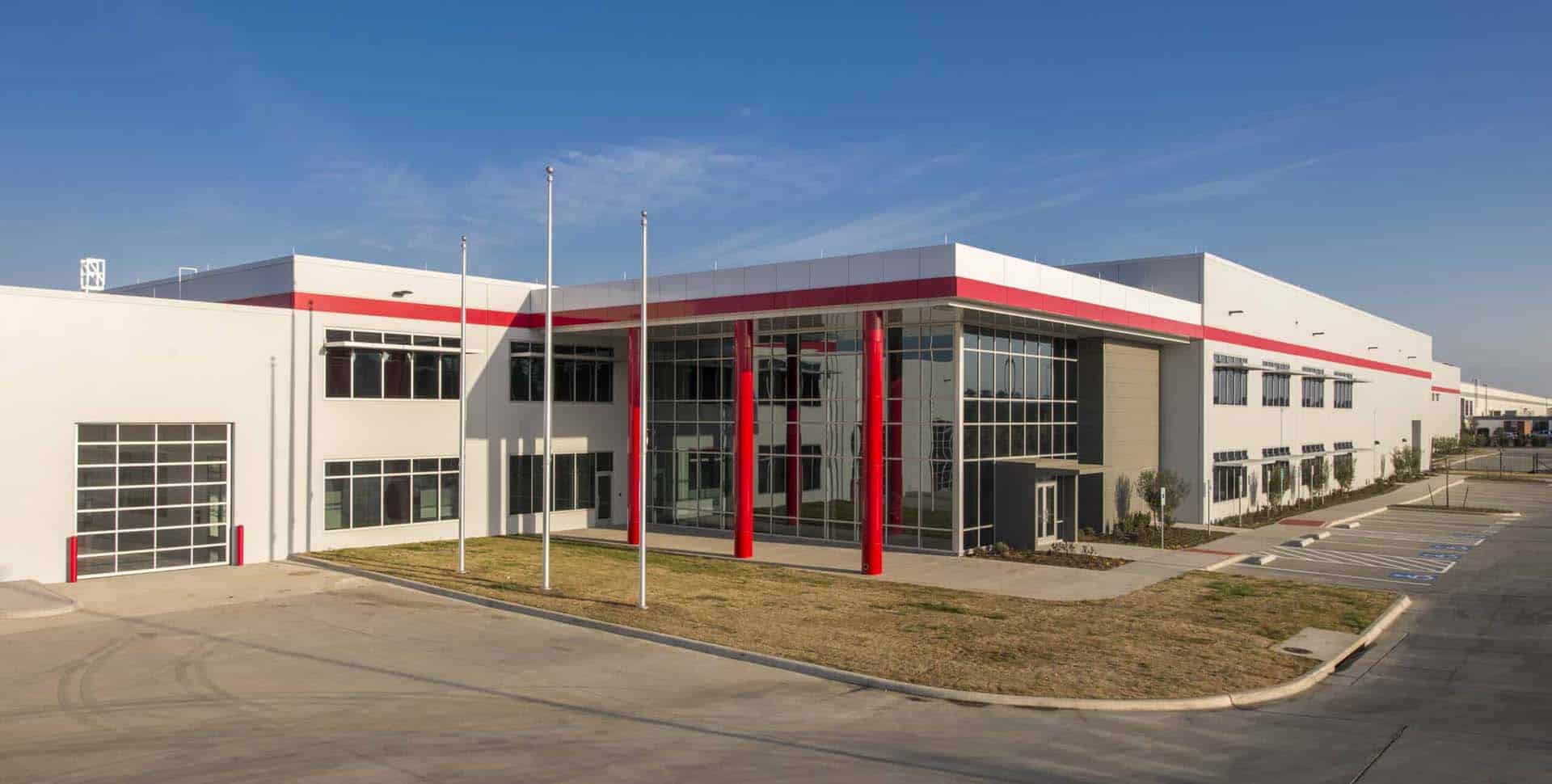 Modern industrial building with large glass entrance, red columns, multiple garage doors, and an adjacent parking lot under a clear blue sky.