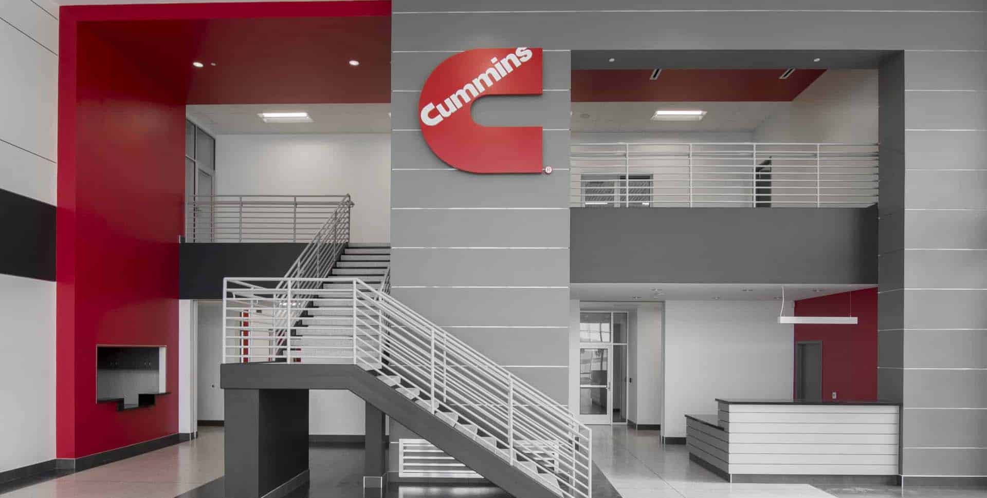Modern office lobby with grey and red accents, a staircase, reception desk, and a large red Cummins logo mounted on the wall.