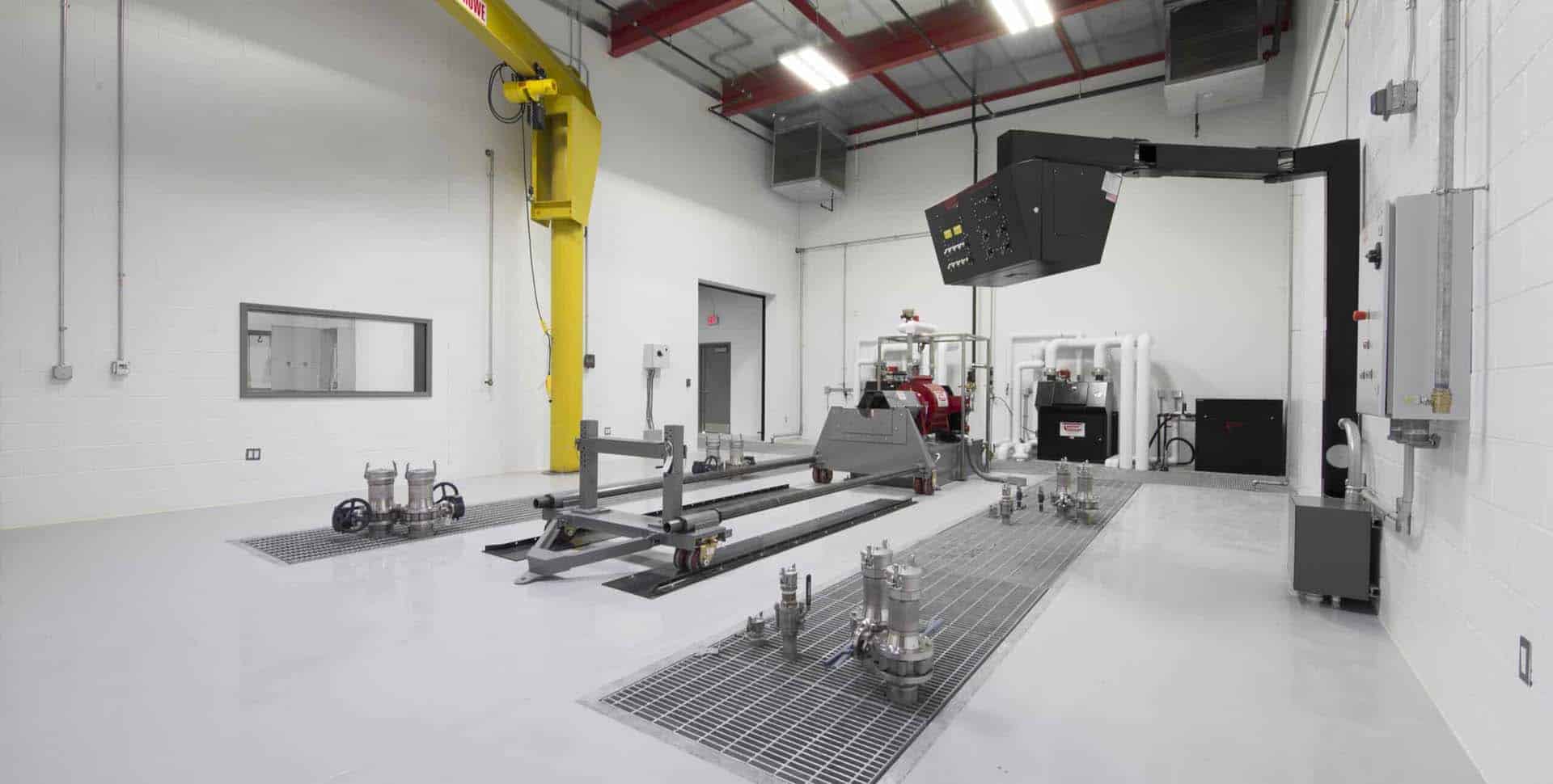 A clean, modern industrial testing facility with metal equipment, ventilation systems, and machinery on a polished white floor under bright overhead lights.