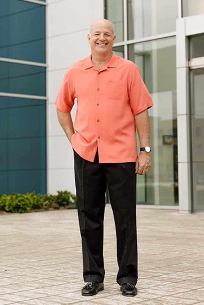 A man stands outside a modern building, wearing a coral short-sleeve shirt, black pants, and black shoes, smiling at the camera.