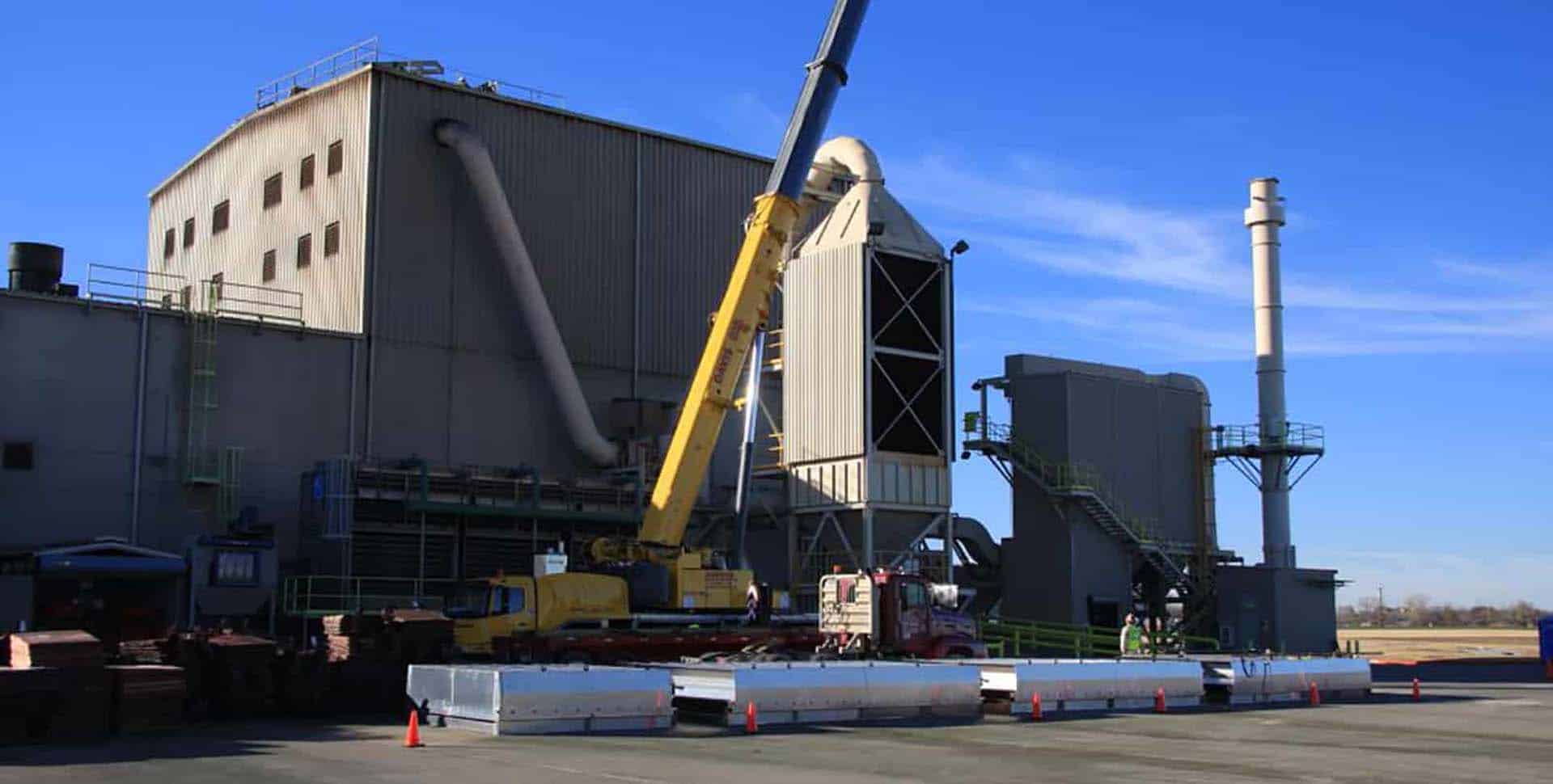 Industrial facility with large metal buildings, a crane, and various equipment under a clear blue sky; barriers and traffic cones in the foreground.