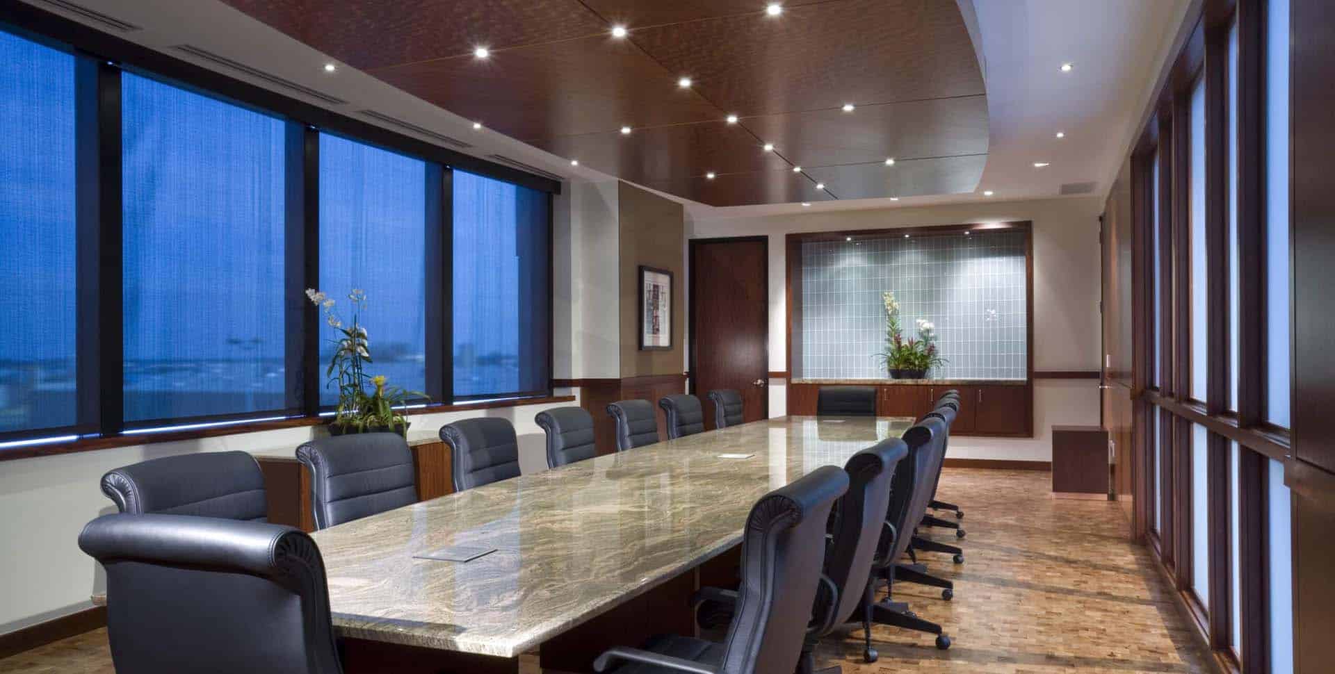 A modern conference room with a long marble table, black leather chairs, wood paneling, large windows, and recessed ceiling lights.
