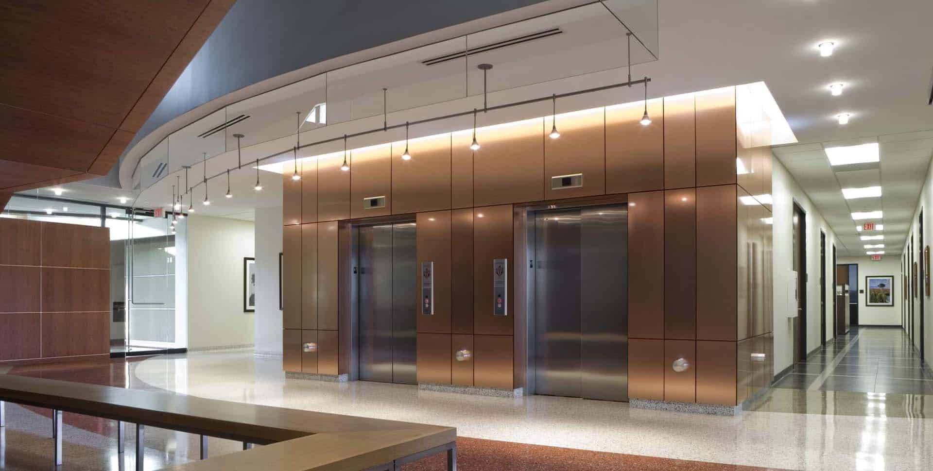 Modern building lobby with two stainless steel elevators set in copper-toned walls, polished floors, overhead pendant lights, and a nearby glass entrance.