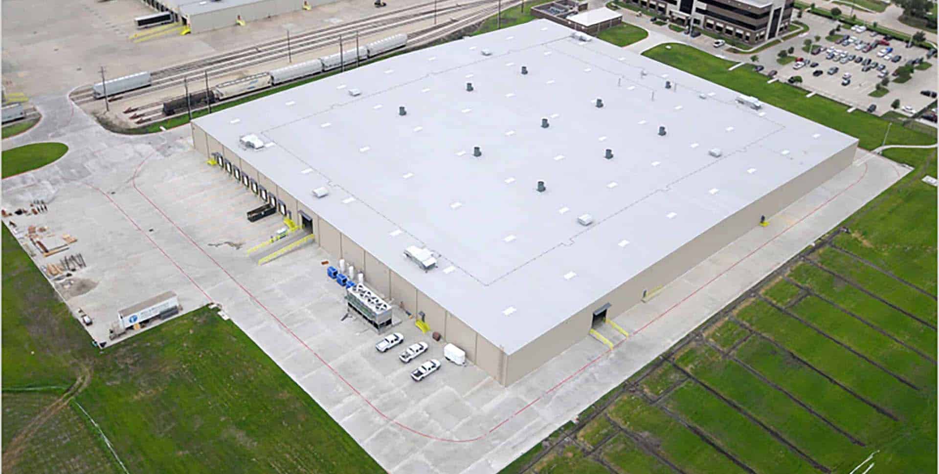 Aerial view of a large industrial warehouse with loading docks, several parked vehicles, and adjacent green spaces.