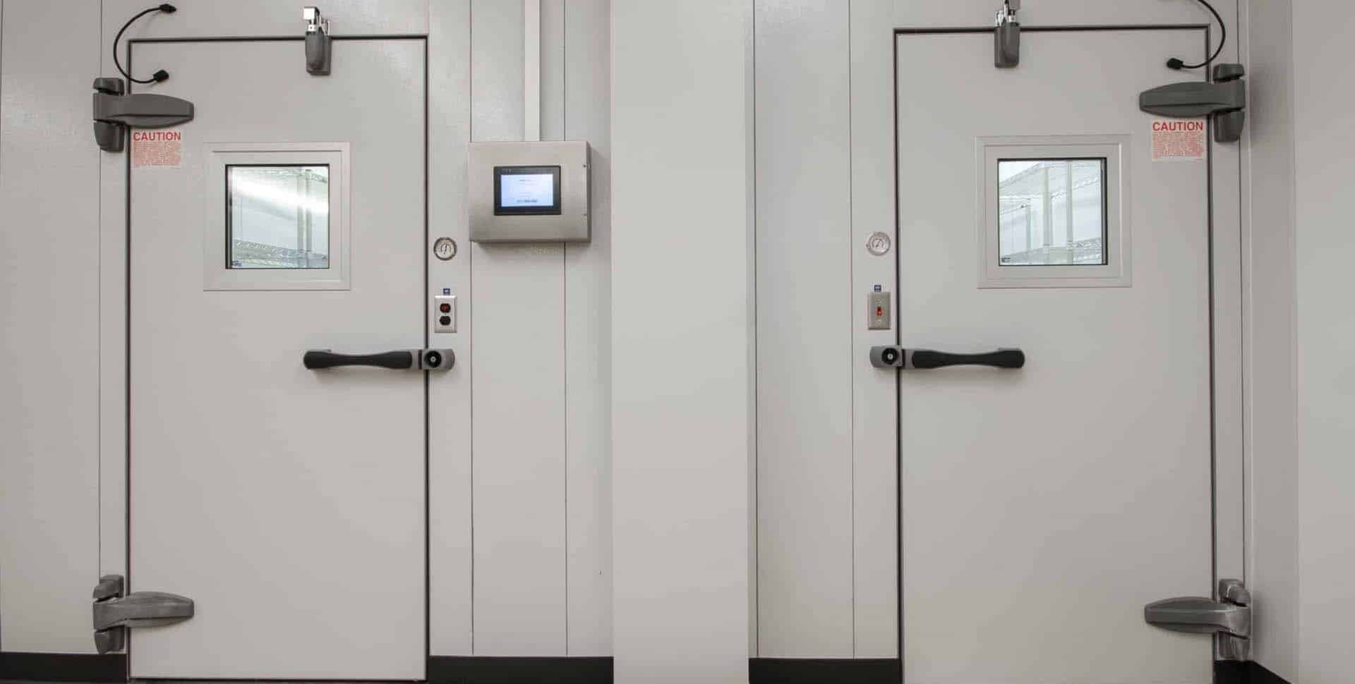 Two industrial white doors with small windows and black handles are set in a white wall; caution signs and a control panel are visible nearby.