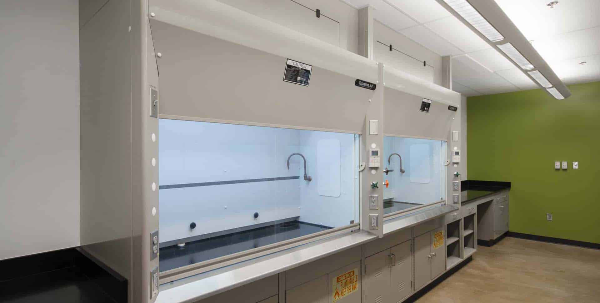 A laboratory with two fume hoods, white cabinetry, black countertops, and a green accent wall. The room is well-lit with fluorescent ceiling lights.