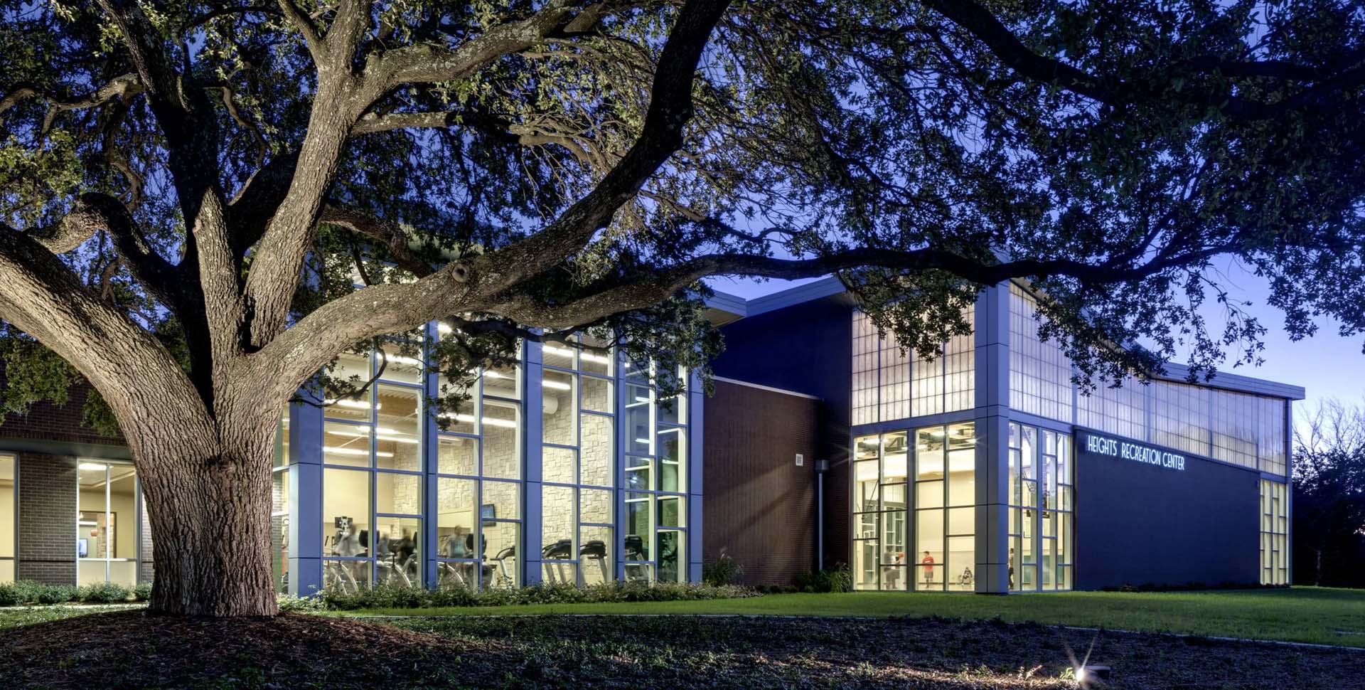 A modern glass-fronted building labeled “Rec & Wellness Center” is illuminated at dusk, with a large tree in the foreground.