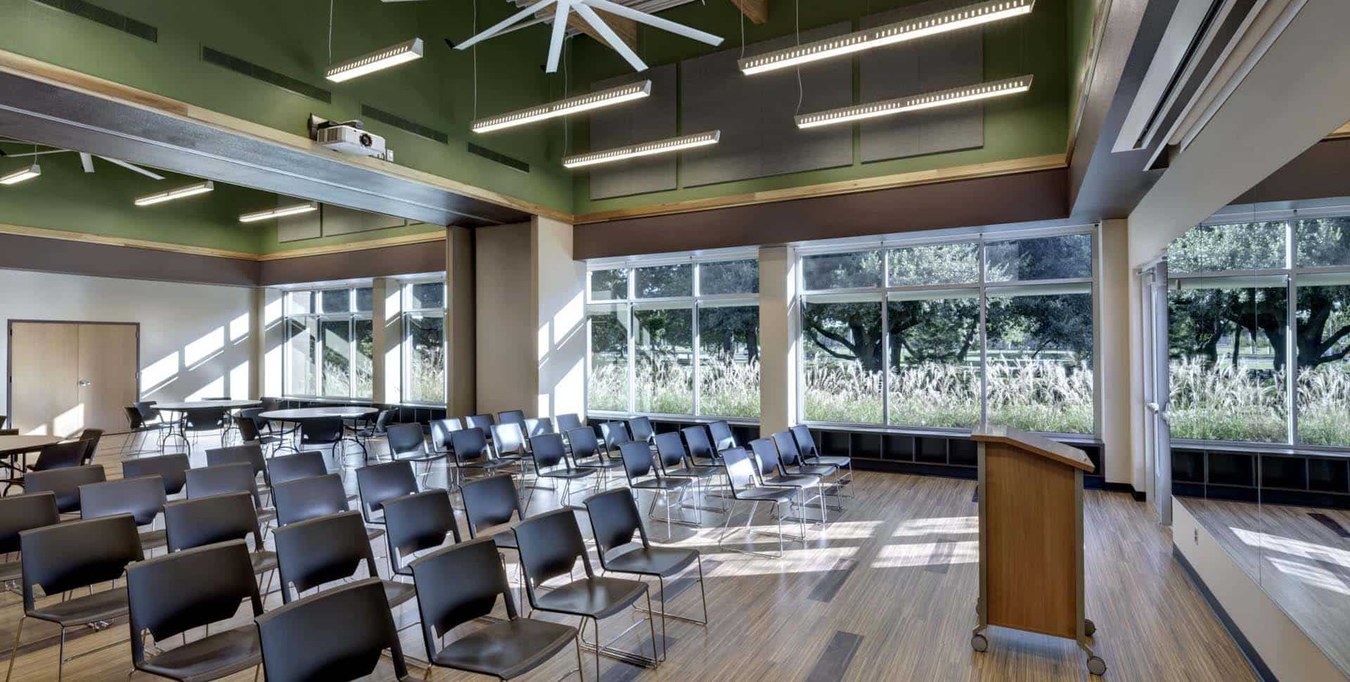 A modern conference room with rows of empty chairs, a podium, large windows with outdoor views, and ceiling lights.