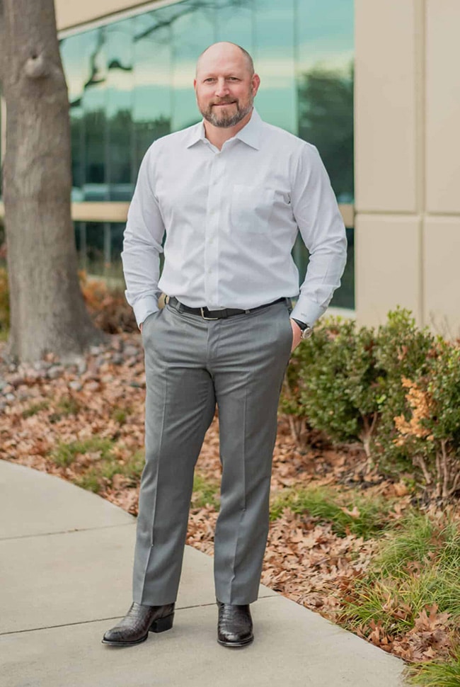 A man with a beard stands outdoors on a sidewalk by a building, wearing a white dress shirt, grey pants, and black shoes, with hands in his pockets.