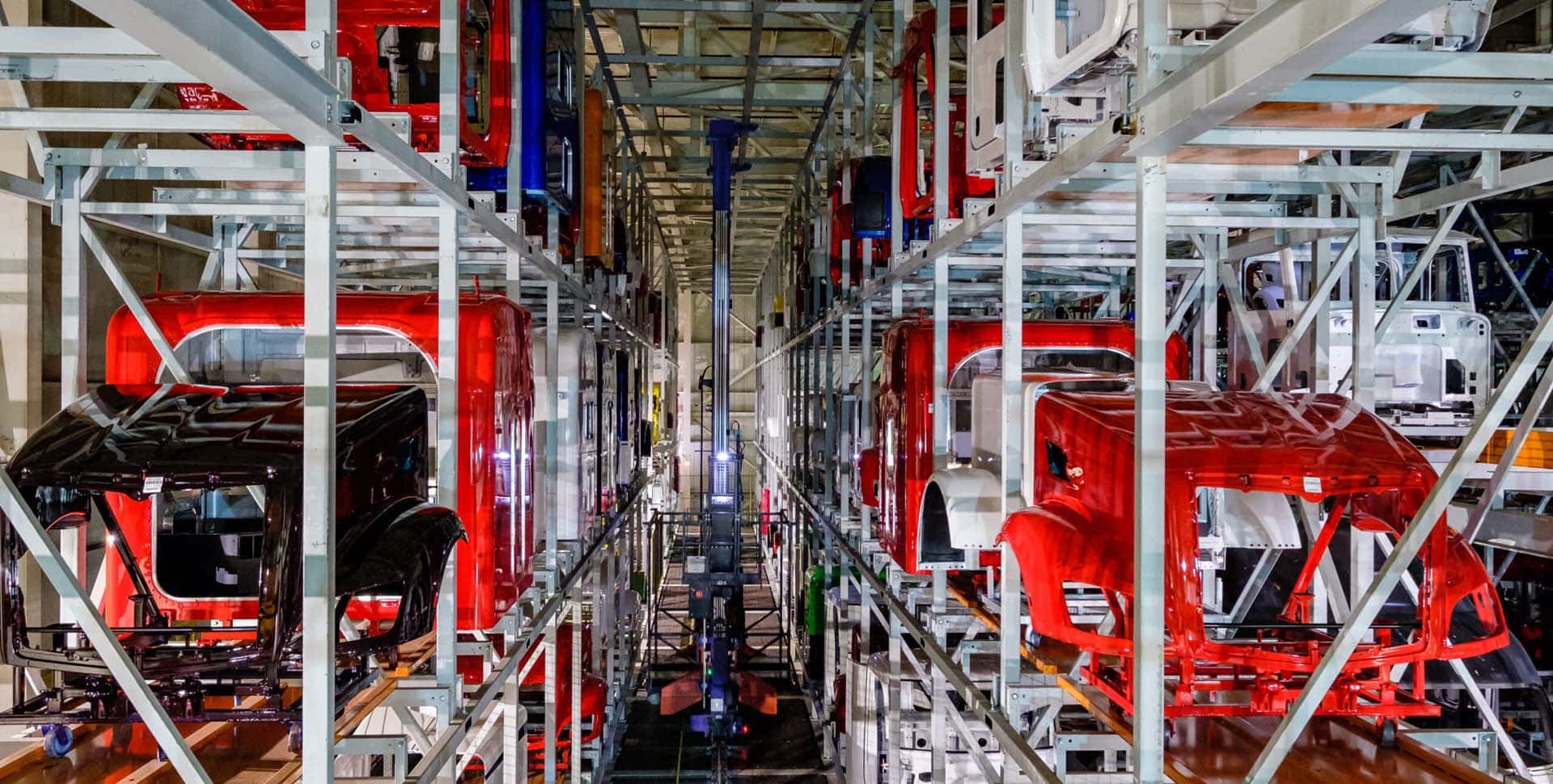 Rows of red and black truck cabs are stored on metal racks in a large, industrial warehouse with automated machinery visible in the center aisle.