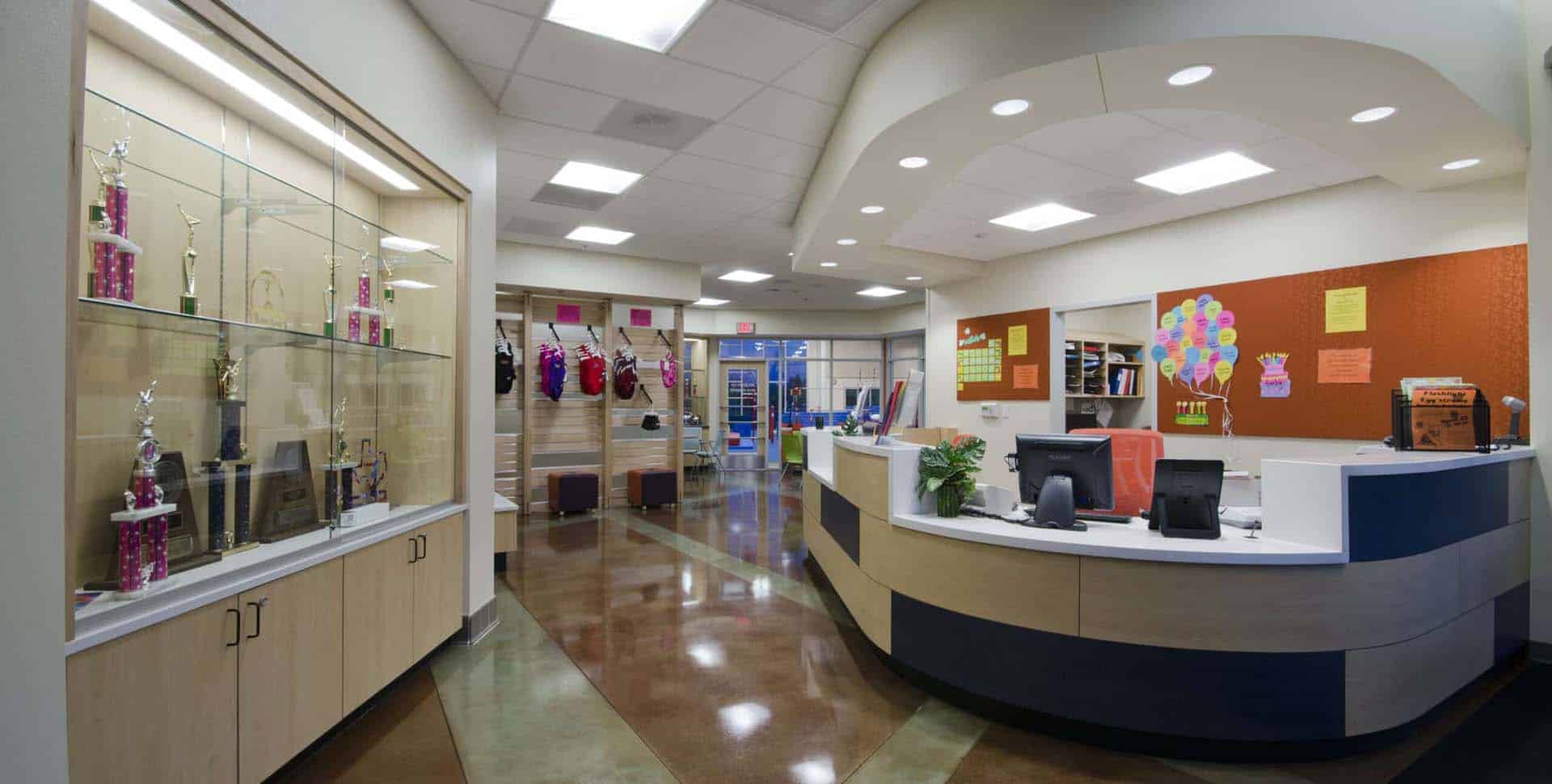 A modern school office with a curved reception desk, computer monitors, trophy display case, bulletin board, and bright overhead lighting.