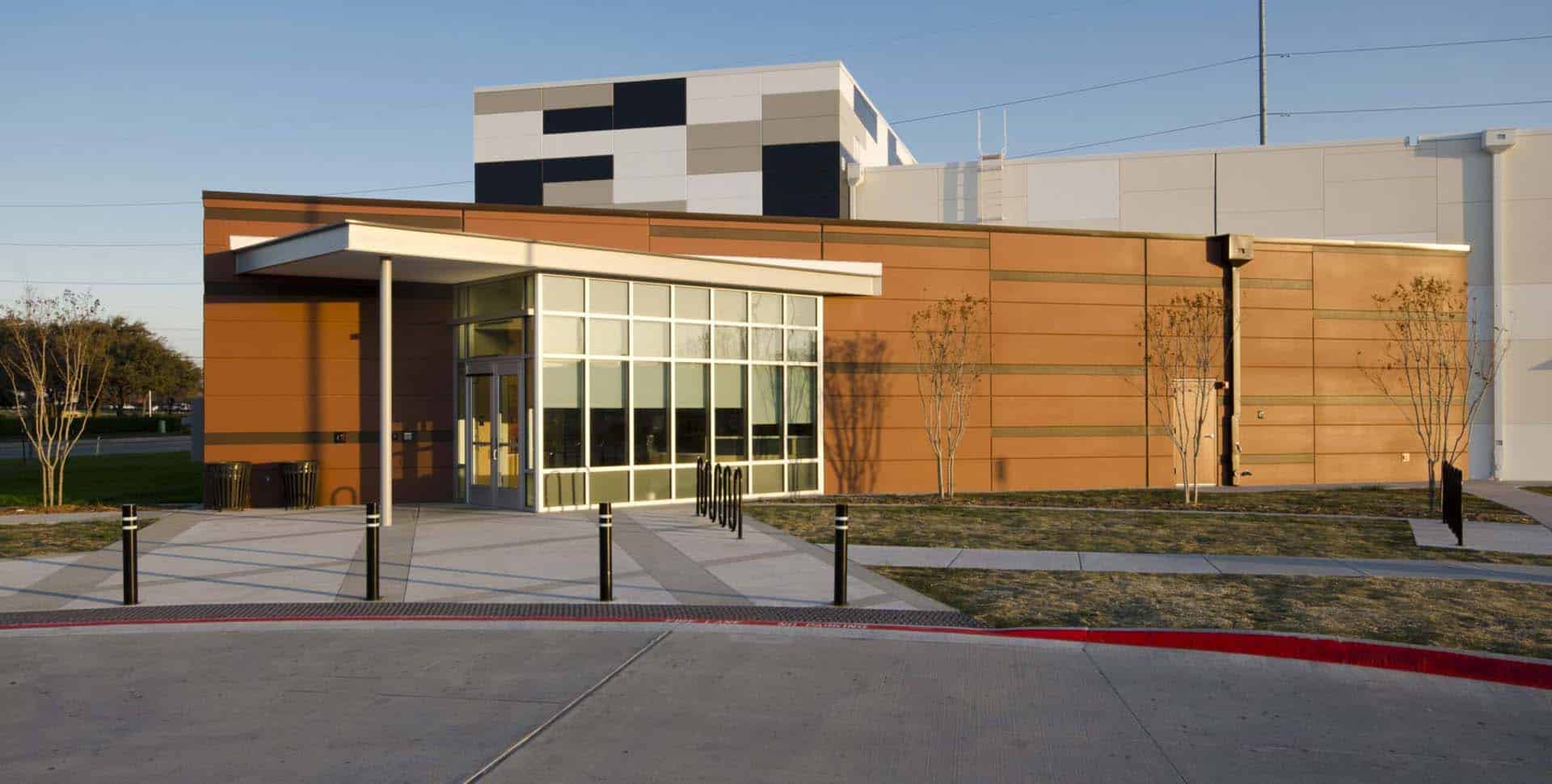 Modern building with orange and white exterior, large windows, and a sidewalk leading to the entrance; sparse landscaping and a few trees in front.
