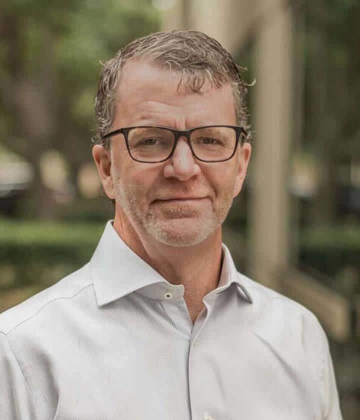 A middle-aged man with glasses and short, graying hair, wearing a light-colored button-up shirt, stands outdoors in front of greenery and a blurred building.