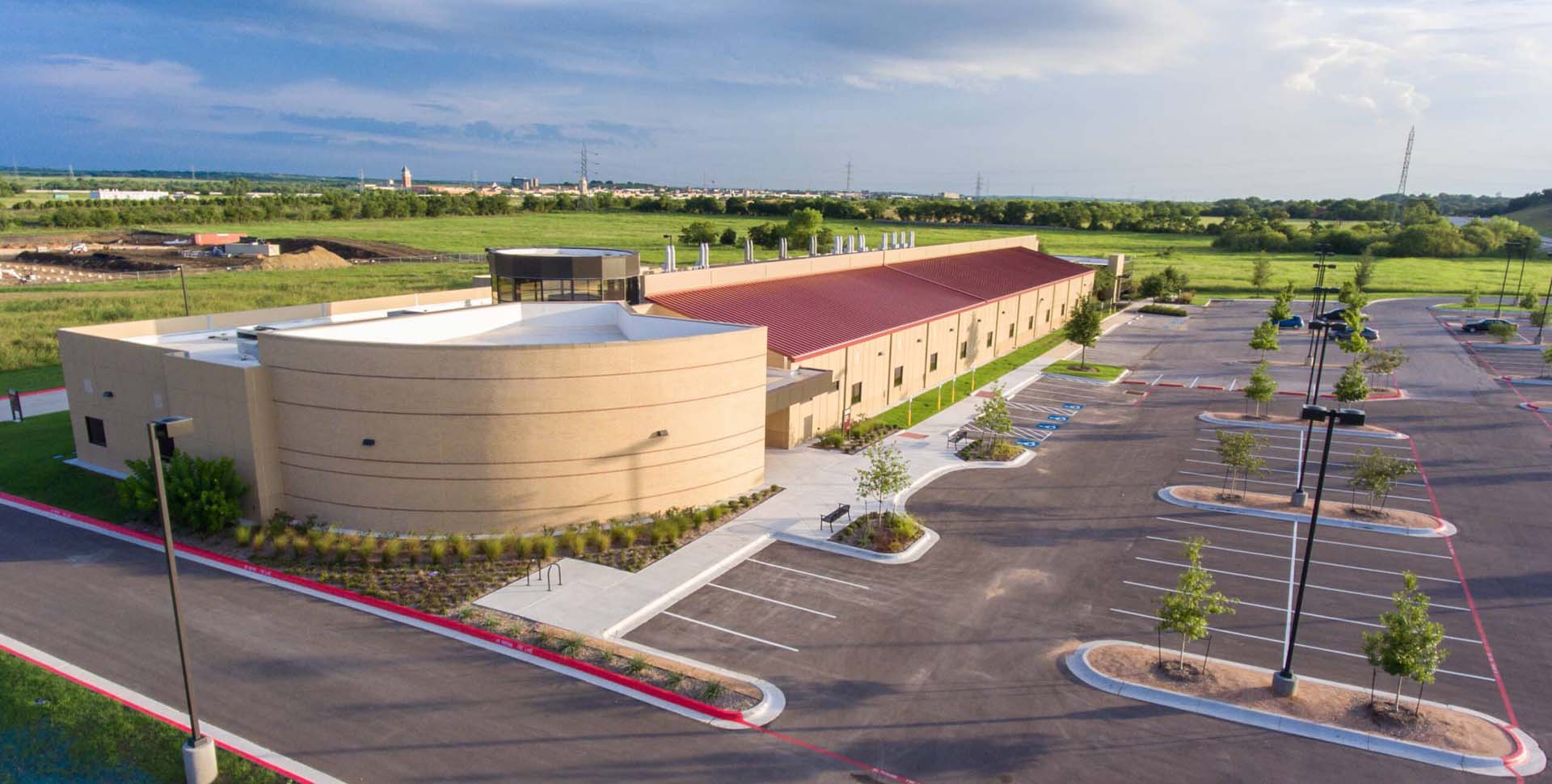 Aerial view of a modern, tan building with a red roof surrounded by parking lots and landscaped greenery, set in a semi-rural area under a partly cloudy sky.