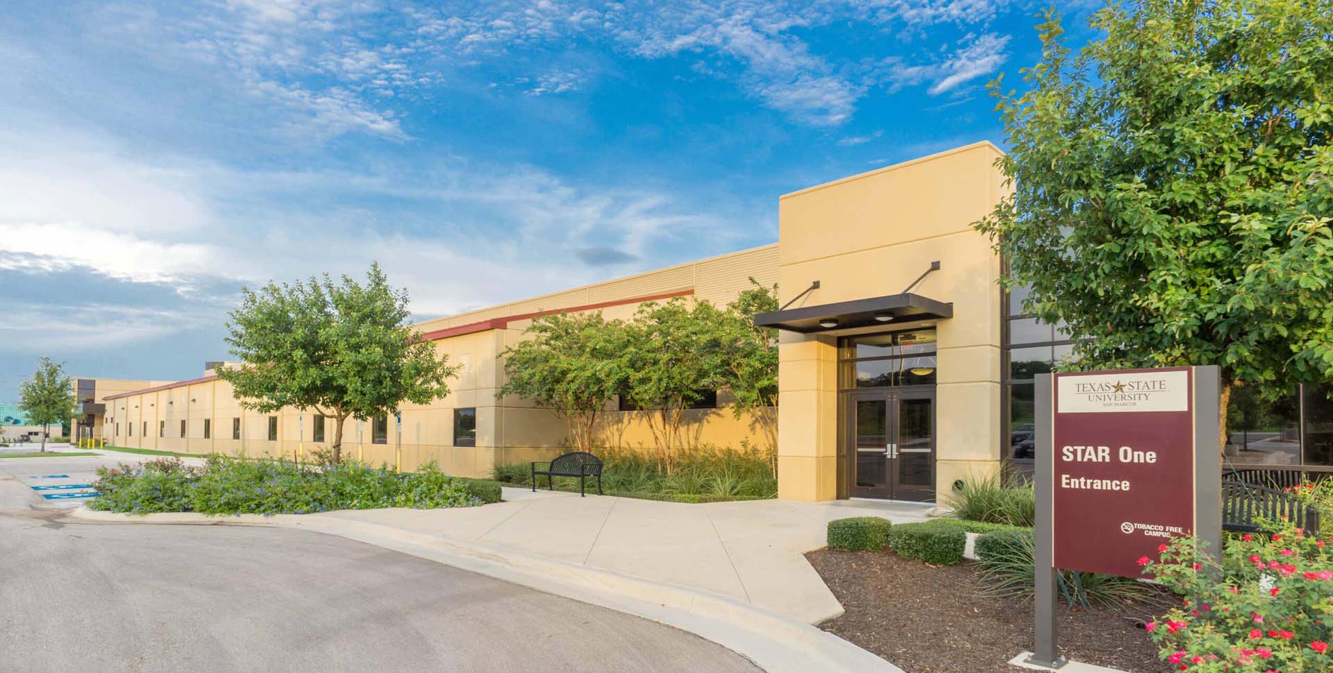 A tan building with large windows and a glass entrance is shown beside a sign reading "Texas State University STAR One Entrance" on a sunny day.