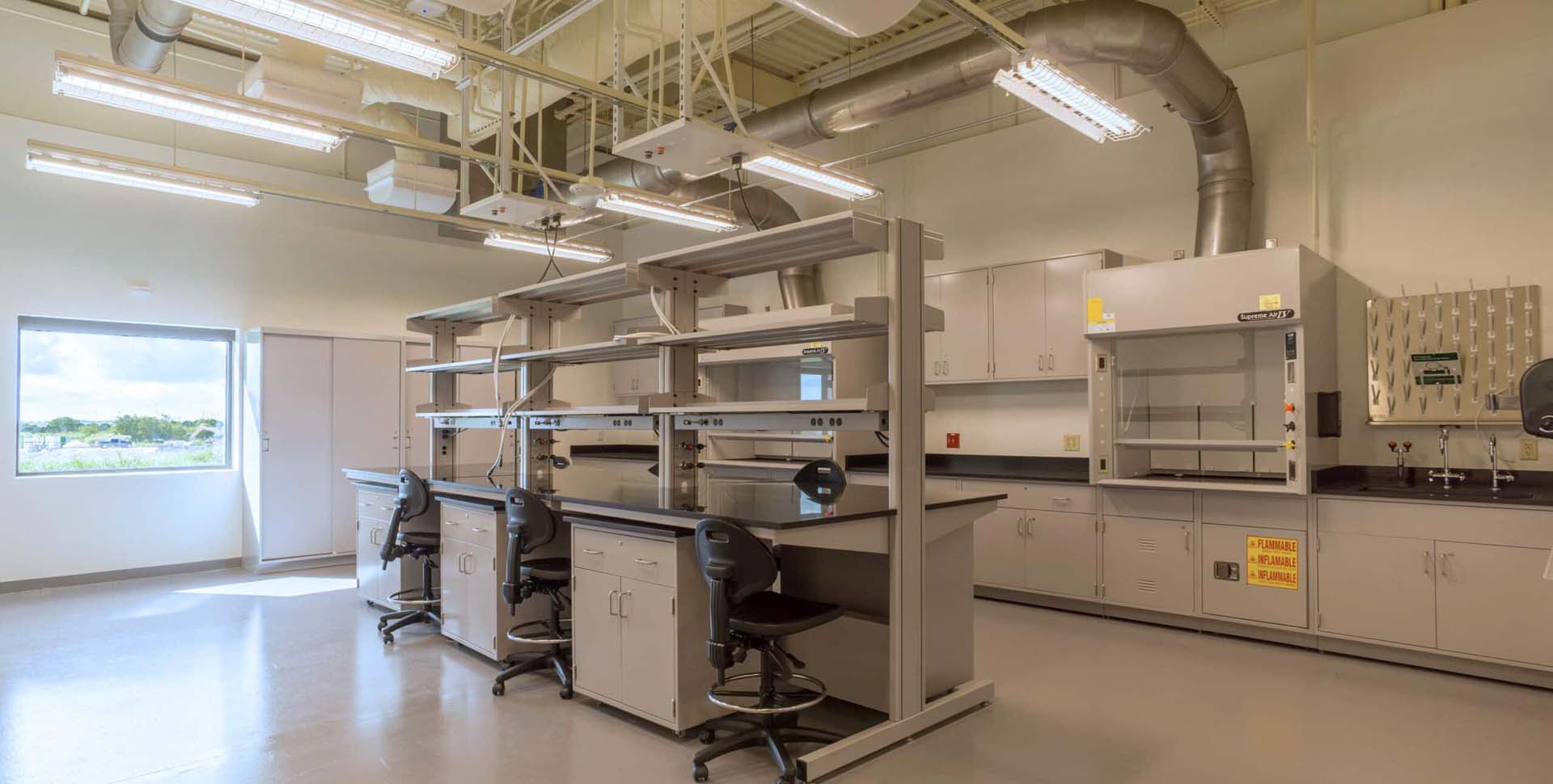 Modern laboratory with empty workstations, shelves, lab chairs, fume hood, cabinets, and overhead lighting; large window provides natural light.