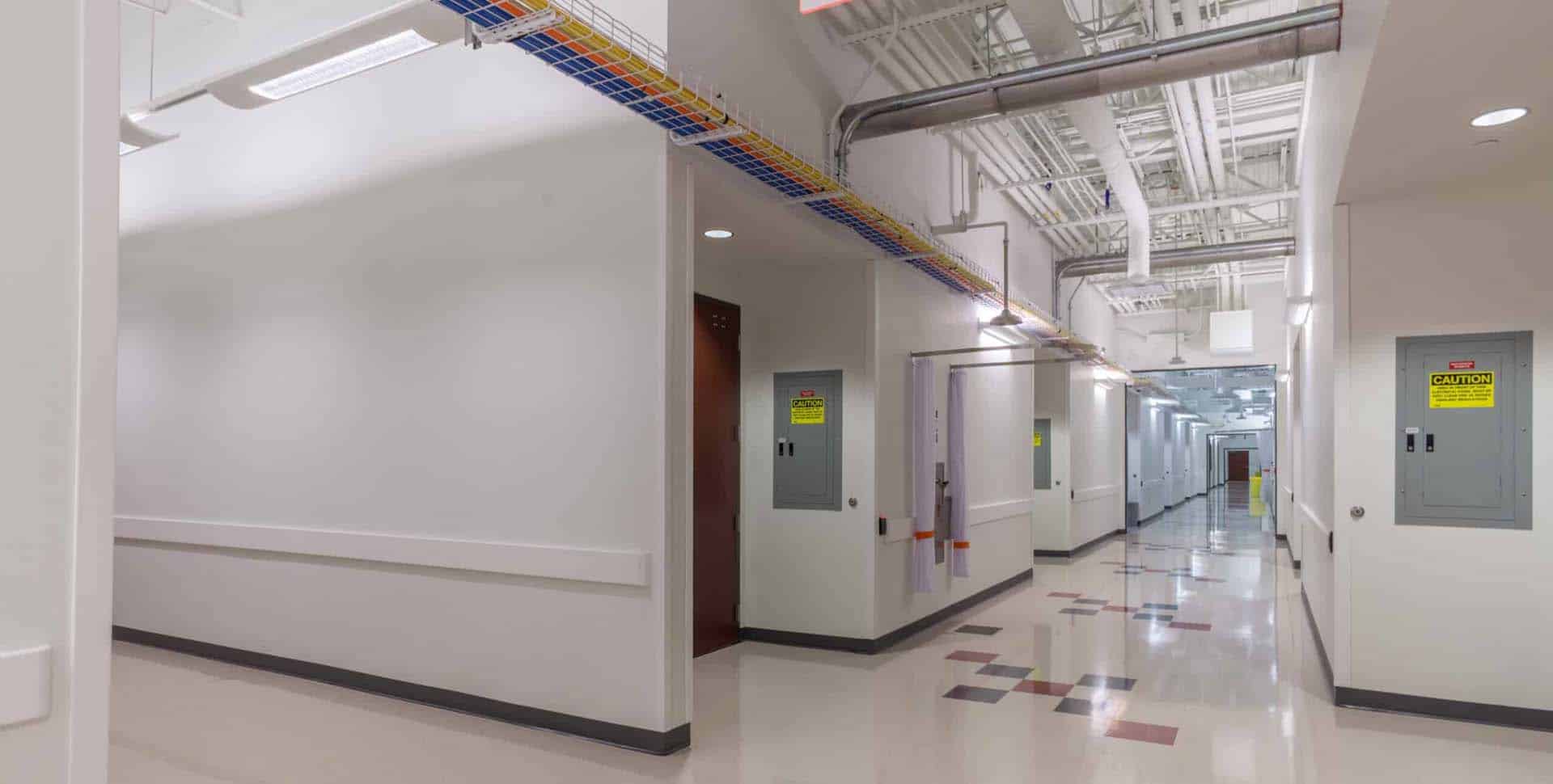 A clean, well-lit hallway in an industrial or institutional building, with electrical panels, exposed pipes, and colored cable trays along the ceiling.