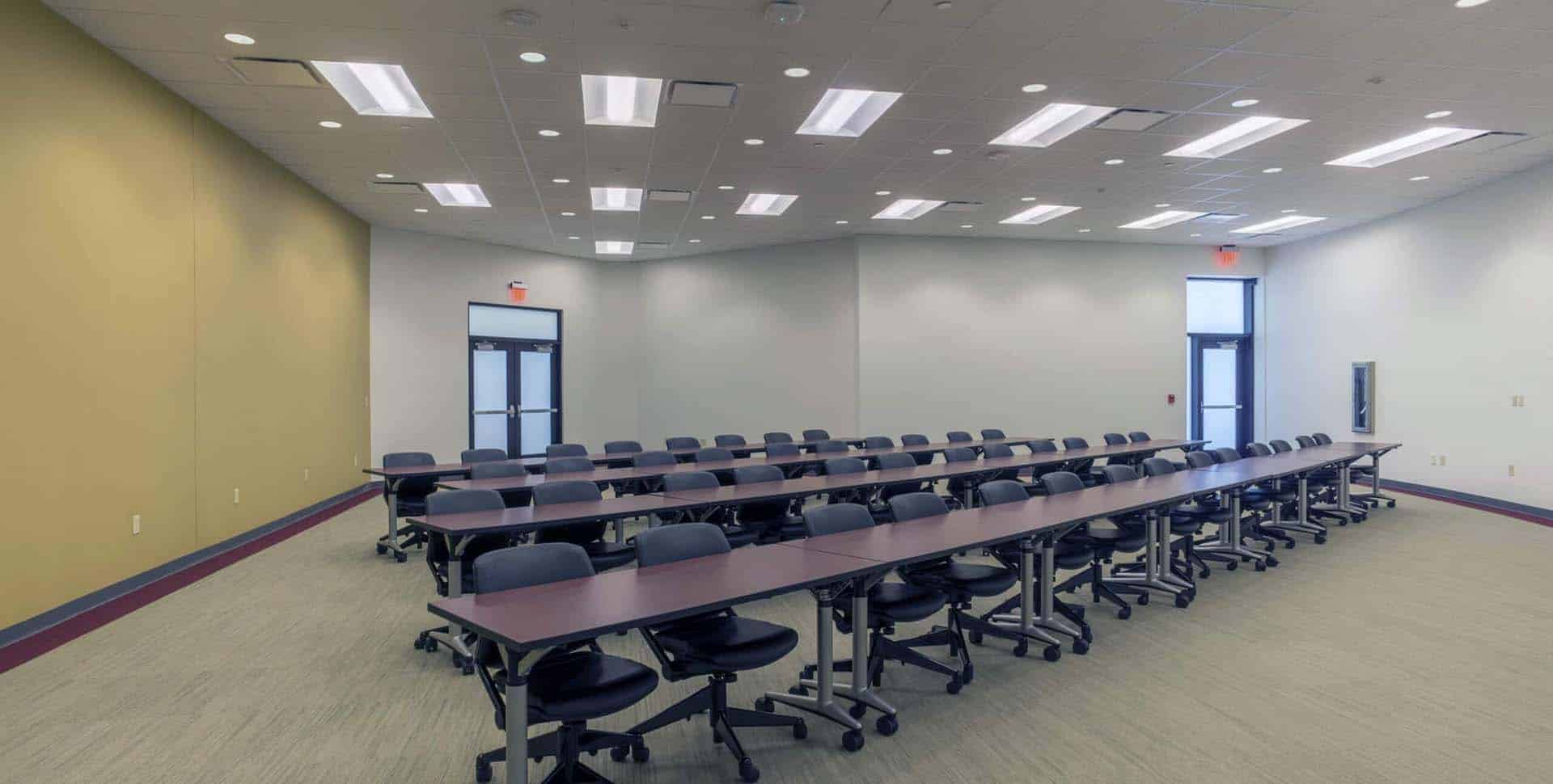 Empty conference room with long tables arranged in a U-shape and office chairs, under fluorescent ceiling lights. The room has beige walls and multiple doors.