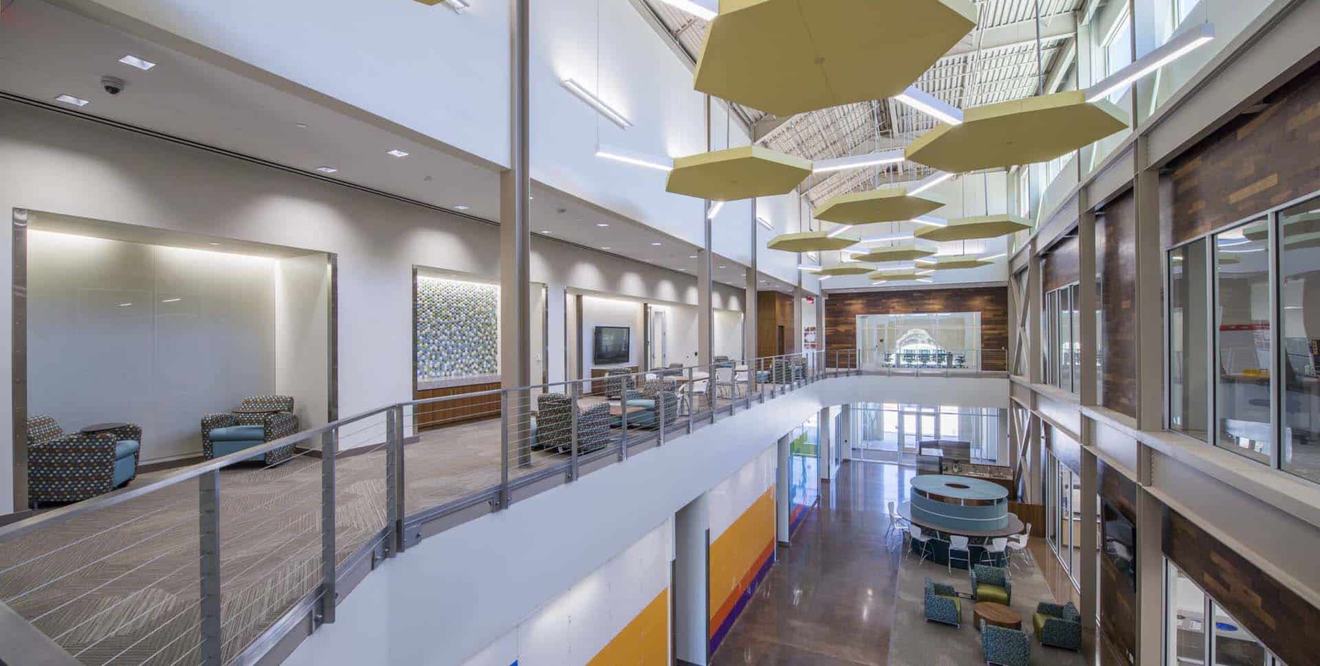 Modern building interior featuring an open mezzanine with glass railings, seating areas, geometric ceiling panels, and large windows providing natural light.